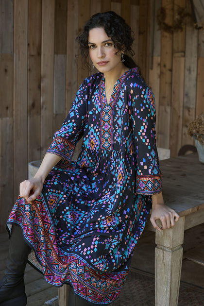 A woman with curly dark hair, dressed in a colorful Geo Kilim Dress, sits on a wooden bench in a rustic setting.