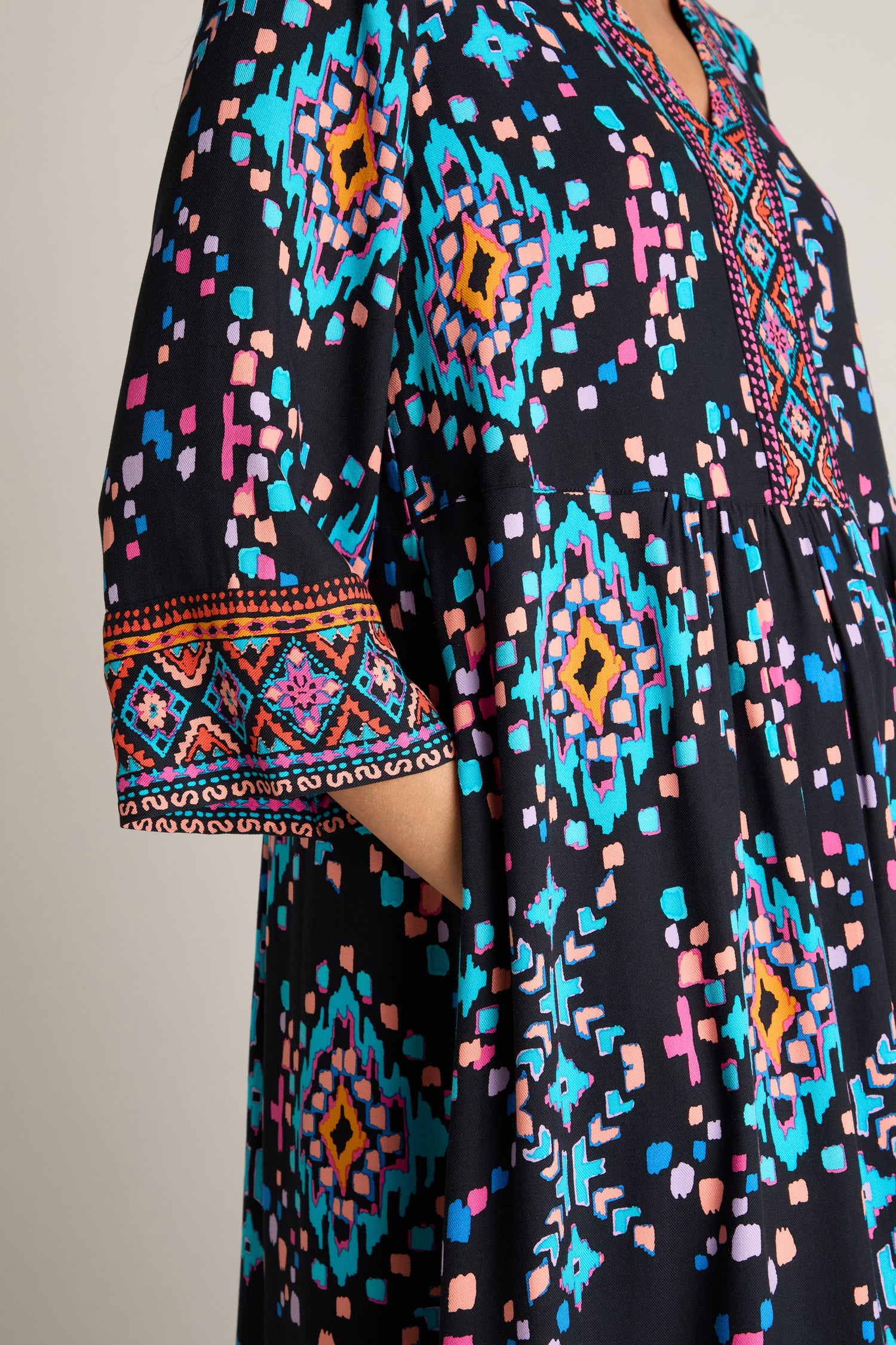 A close-up shot shows someone donning the Geo Kilim Dress in black, featuring vibrant geometrical patterns in blue, pink, and orange. The sleeve is embellished with detailed embroidery.