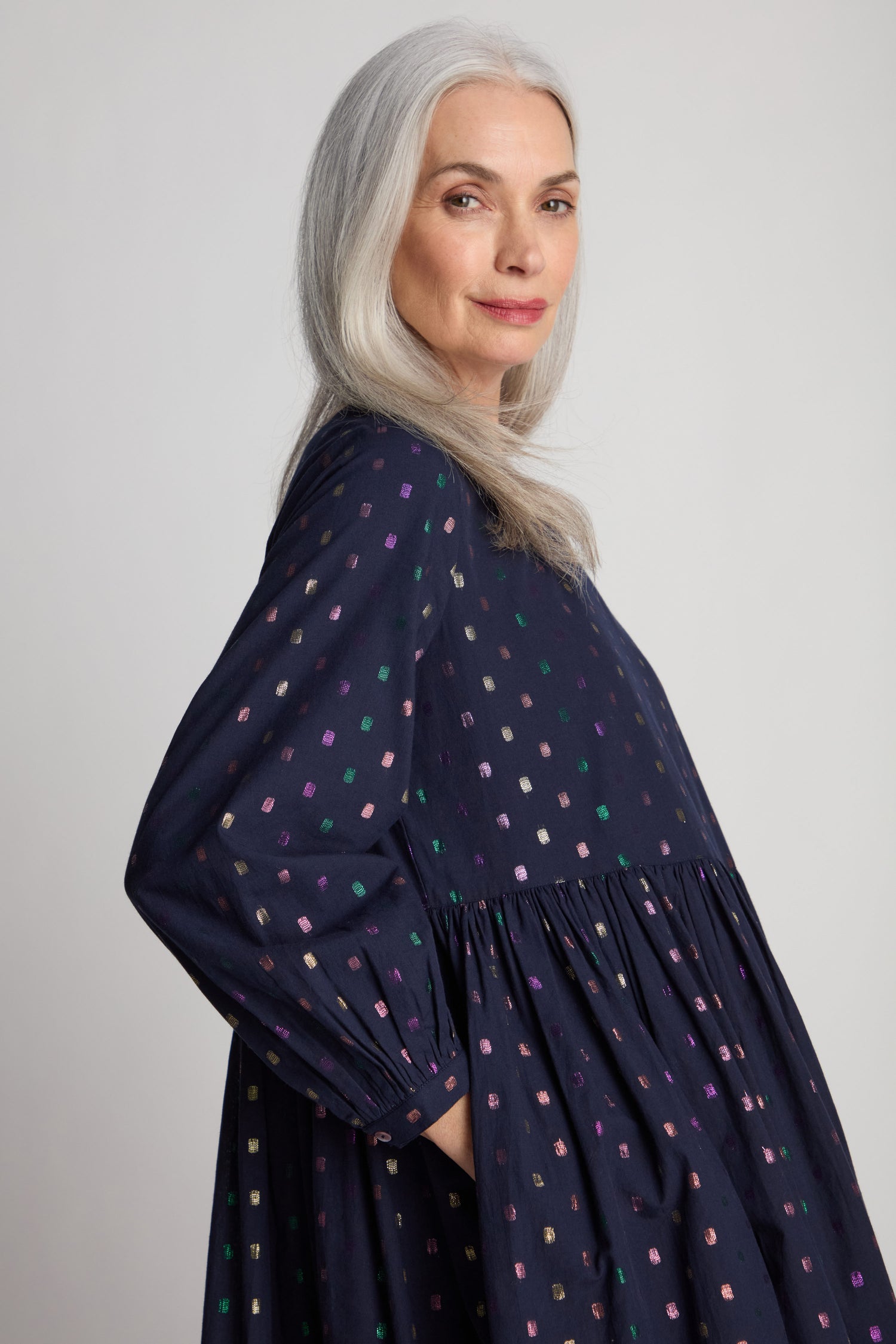 An older woman with long gray hair stands in profile, wearing the Multi Square Lurex Pleated Dress featuring a dotted pattern and colorful sparkle details. She has a slight smile as she looks at the camera, hands in her pockets.