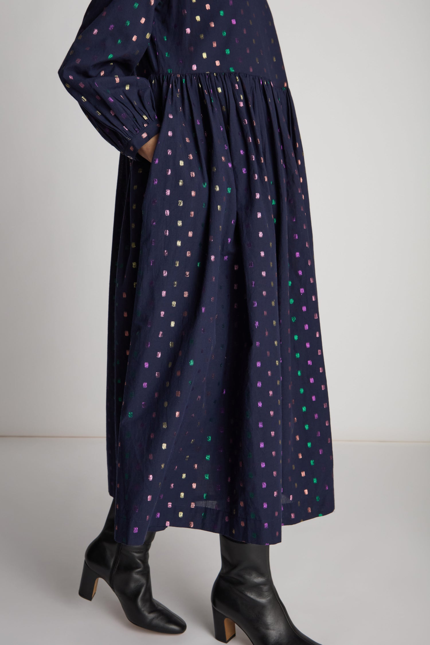 A person stands against a plain background, wearing the knee-length, long-sleeved Multi Square Lurex Pleated Dress paired with black boots, showcasing its voluminous silhouette and colorful sparkle details.