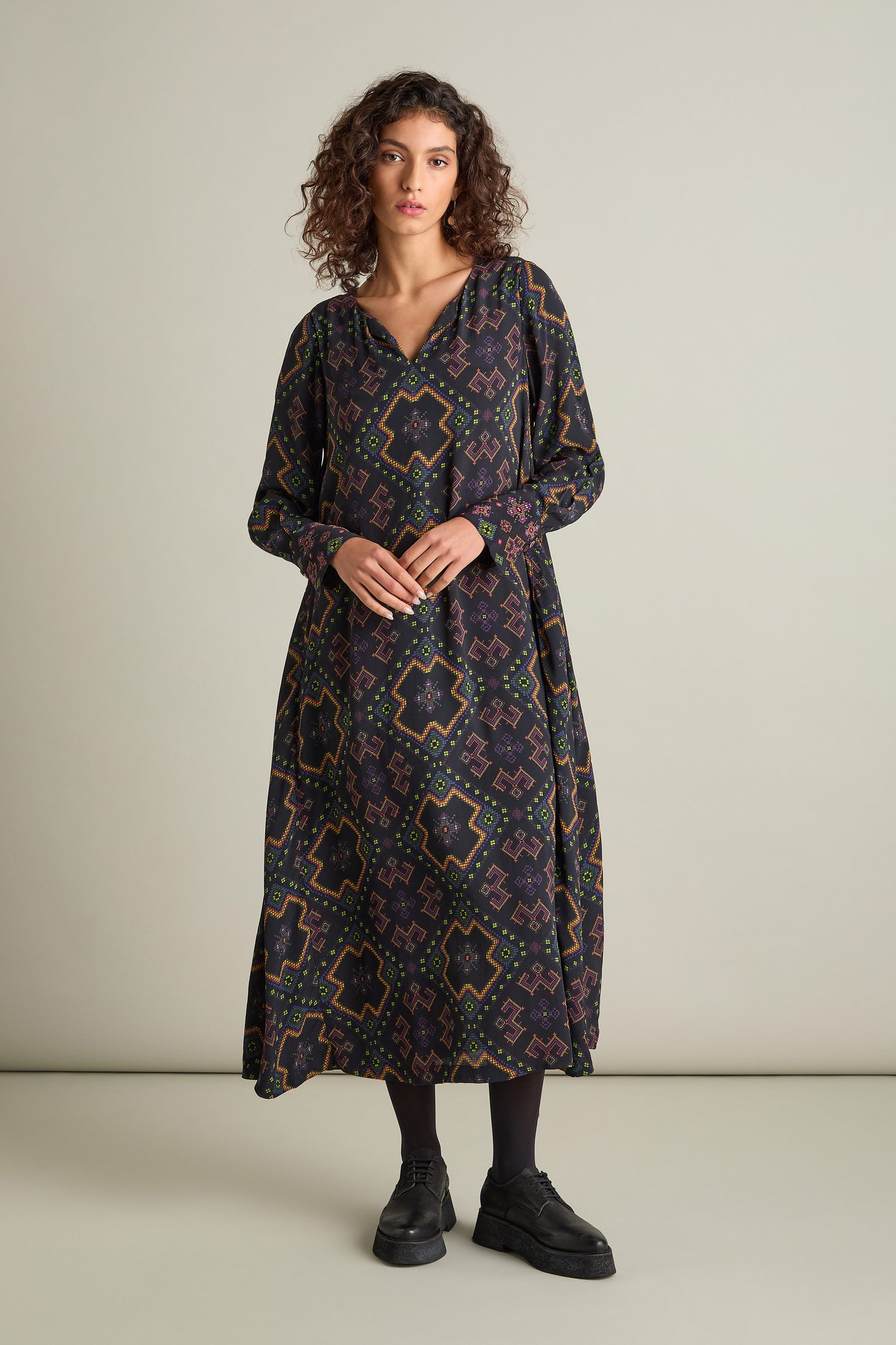 A person stands against a plain background wearing the Pixelated Jacquard Print Dress, a long and stylish garment adorned with an intricate pixelated pattern. They have curly hair and are sporting black shoes.