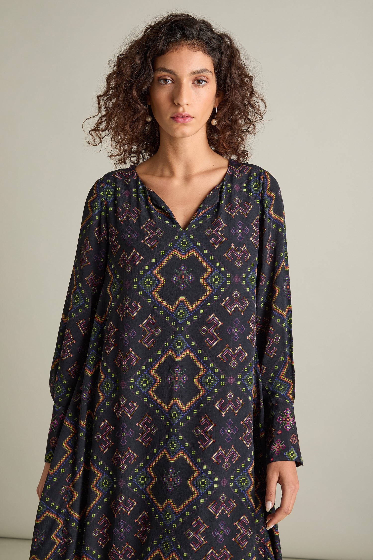 A person with curly hair, wearing the Pixelated Jacquard Print Dress—a long-sleeved black garment adorned with a colorful geometric print—stands against a neutral background.