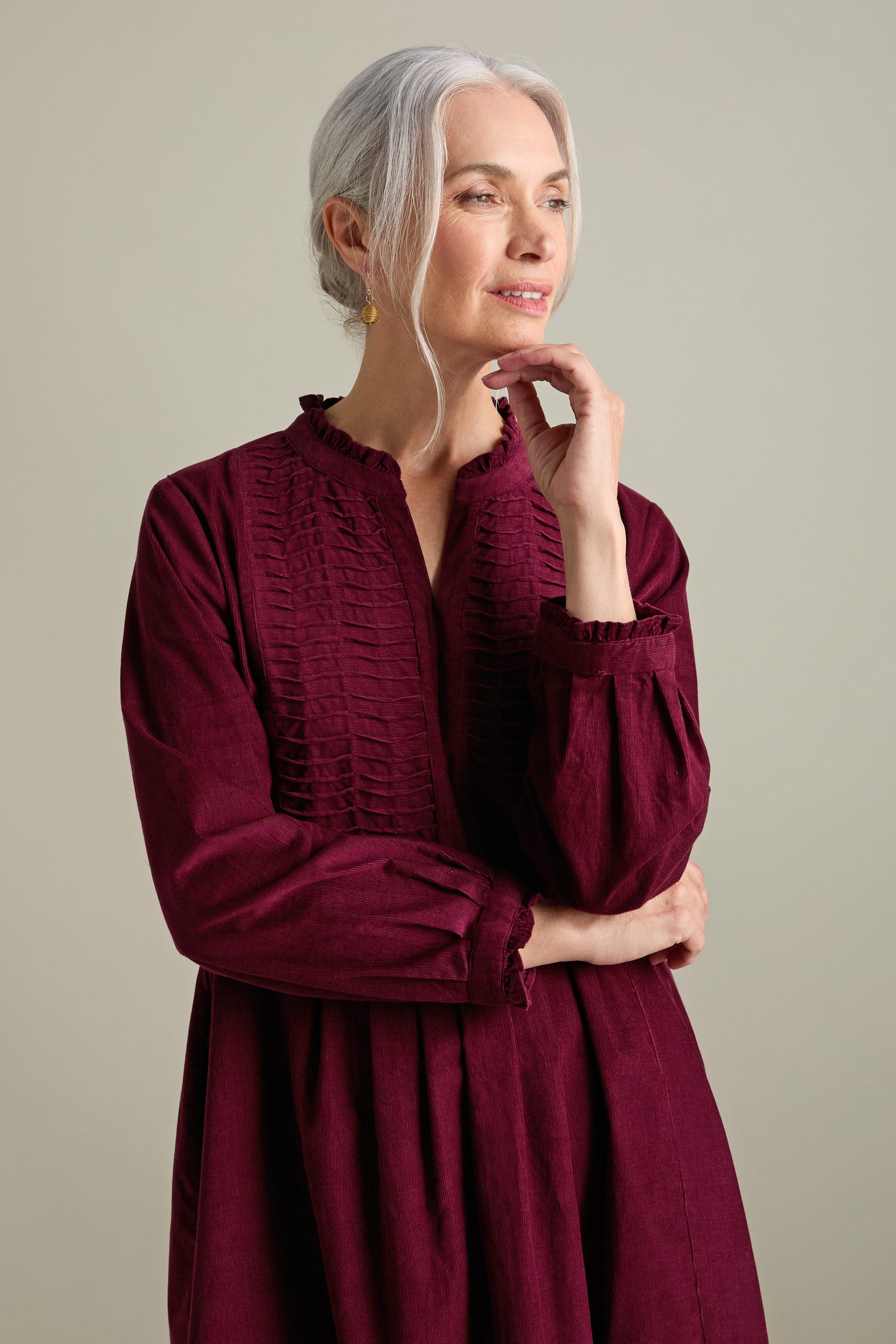 A woman with gray hair in a braid is wearing a long-sleeved, burgundy Pinwale Cord Pleat Front Dress. She is looking off to the side with her hand resting on her chin.