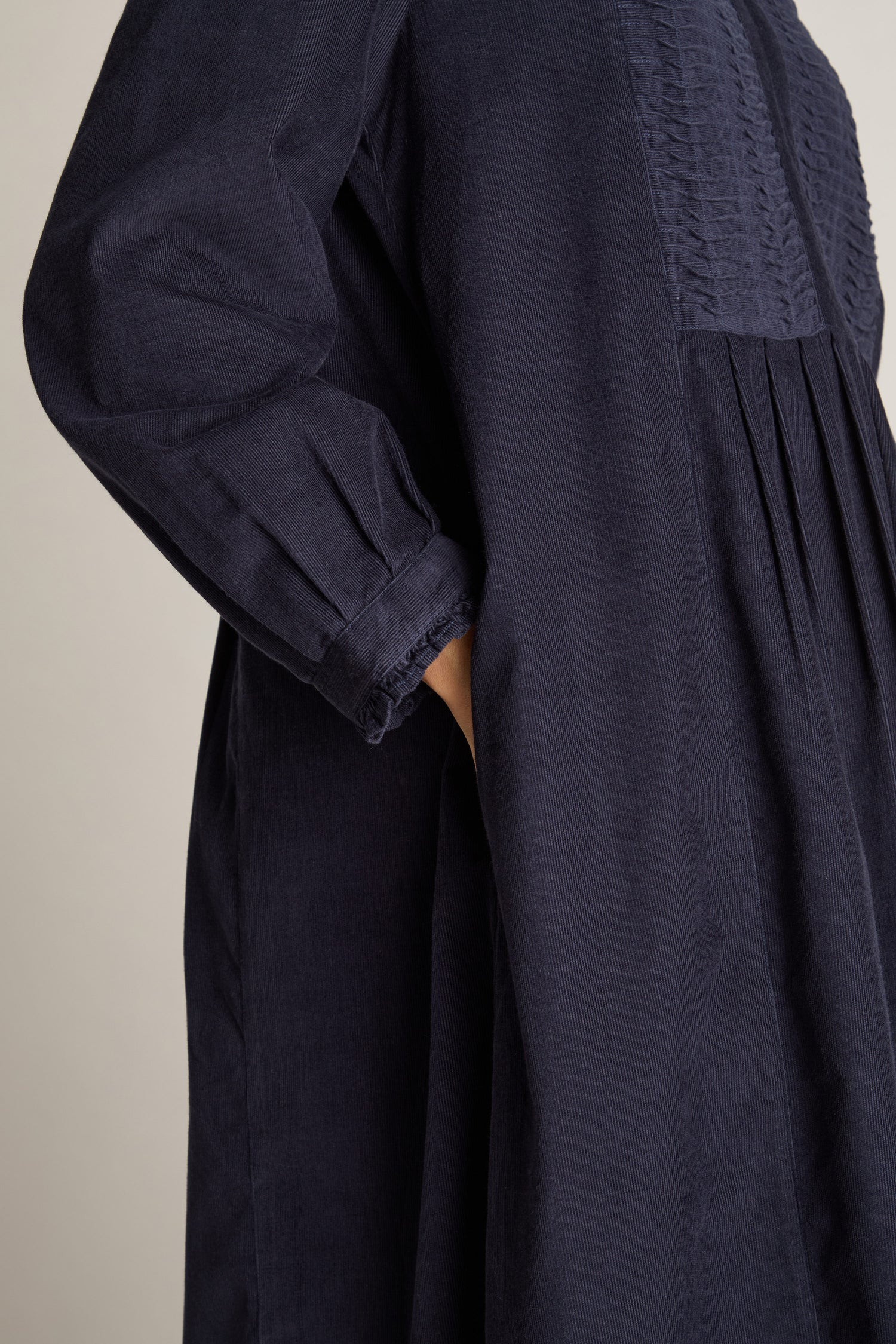 A person is wearing a dark, long-sleeved Pinwale Cord Pleat Front Dress featuring an A-line silhouette and pintuck details, with their hand tucked into a side pocket.