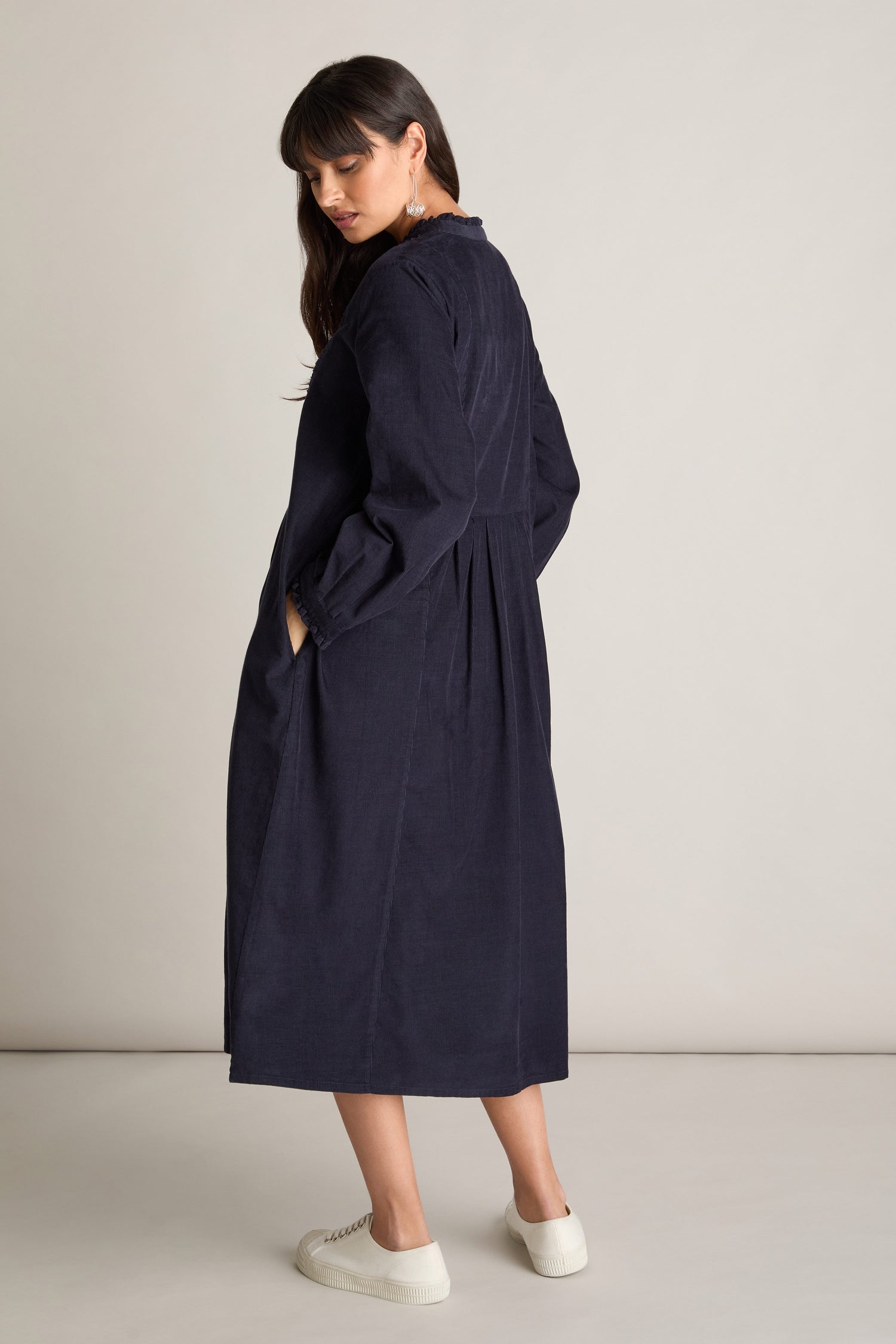 Person wearing a Pinwale Cord Pleat Front Dress in navy blue with long sleeves and white sneakers, standing and facing slightly away from the camera.