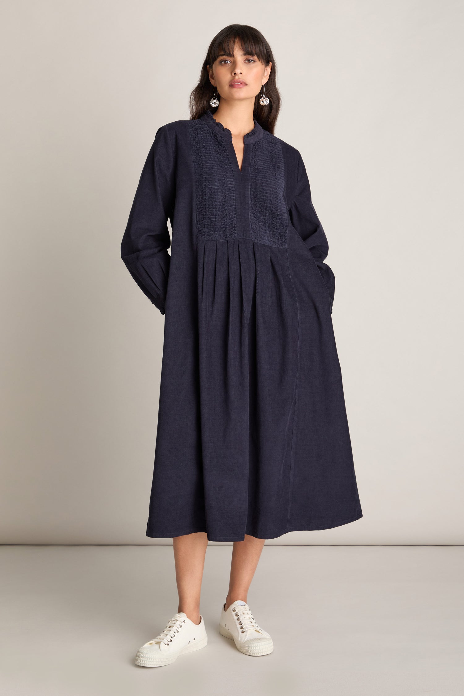 A woman wearing the Pinwale Cord Pleat Front Dress, a long-sleeve, dark blue midi dress with an A-line silhouette and a V-neckline, is paired with white sneakers and standing against a plain background.