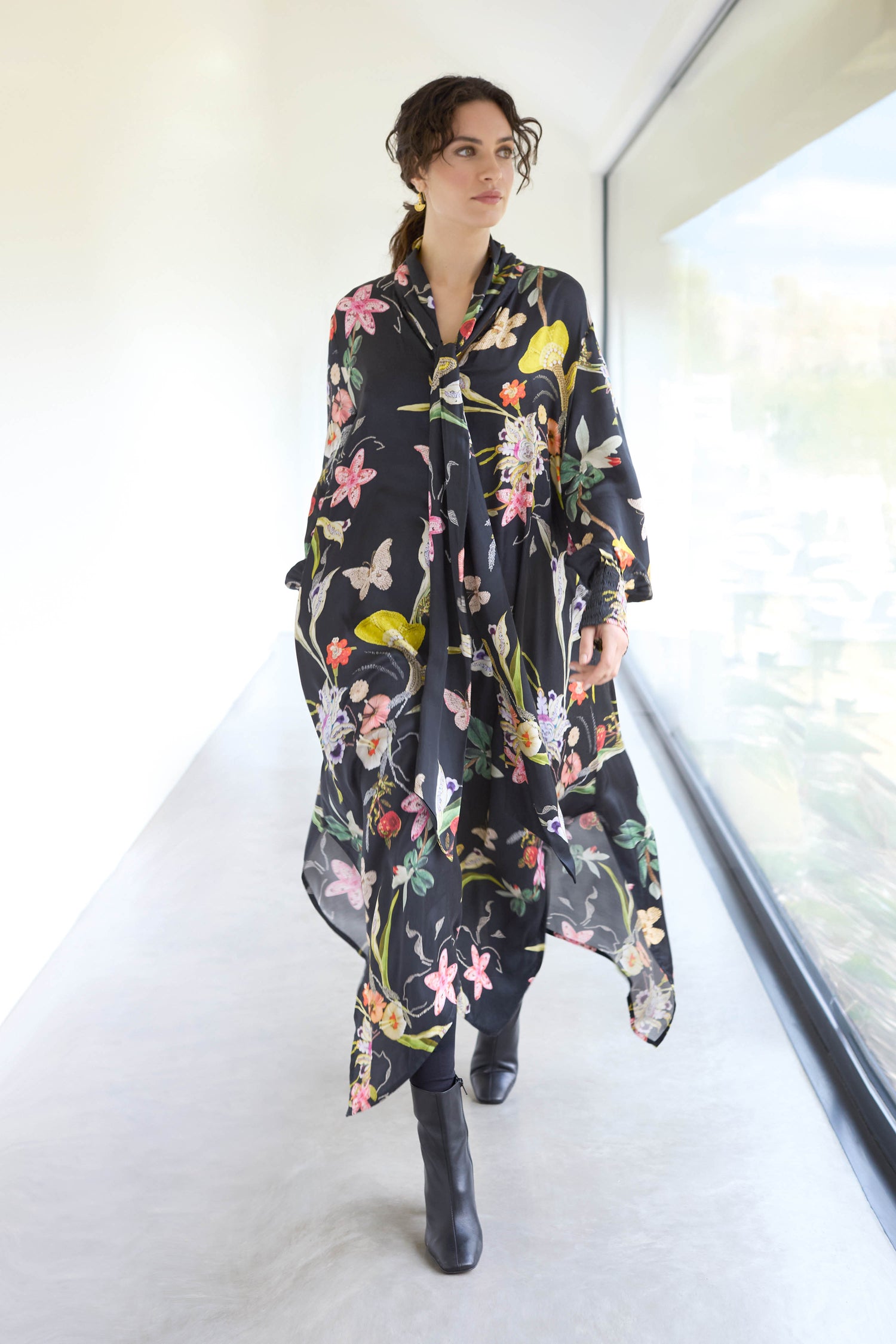 A person strolls down a bright hallway, wearing a Vintage Floral Tie Front Dress with an A-line silhouette, paired perfectly with black boots.