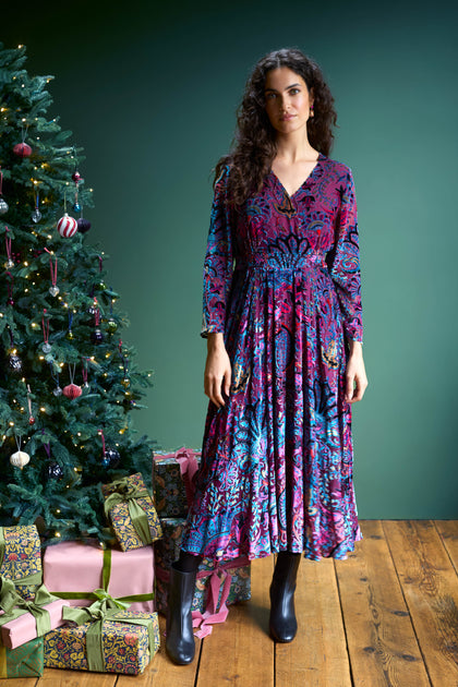 A woman in a Winter Paisley Devore Flared Dress stands next to the GRD6069-WPD decorated Christmas tree, with gifts tucked underneath on a wooden floor.