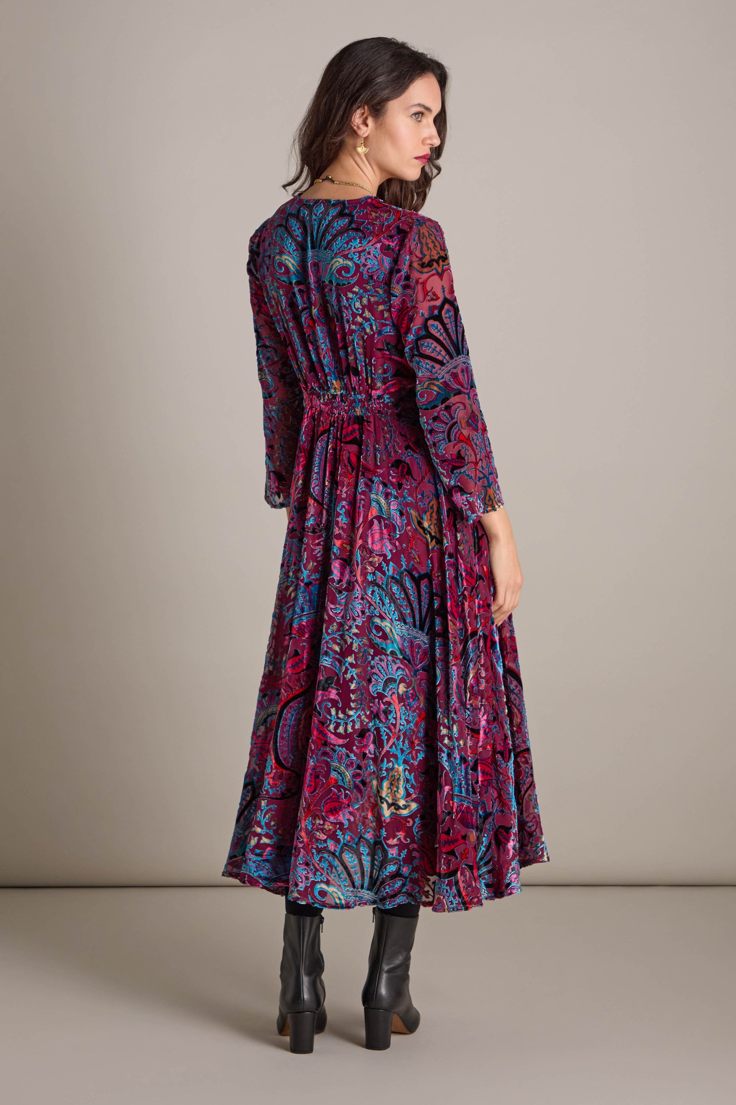 Someone in a colorful Winter Paisley Devore Flared Dress is facing away from the camera, paired with black boots.