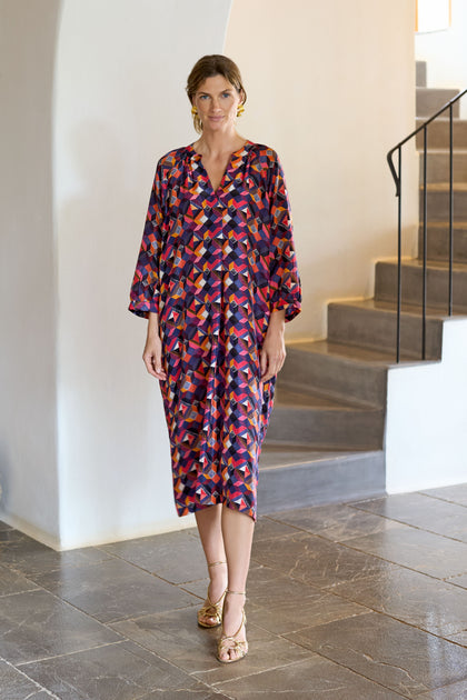 A person in a Small Stitched Squares Silk Dress with a colorful geometric design stands inside near a staircase, maintaining a neutral expression.