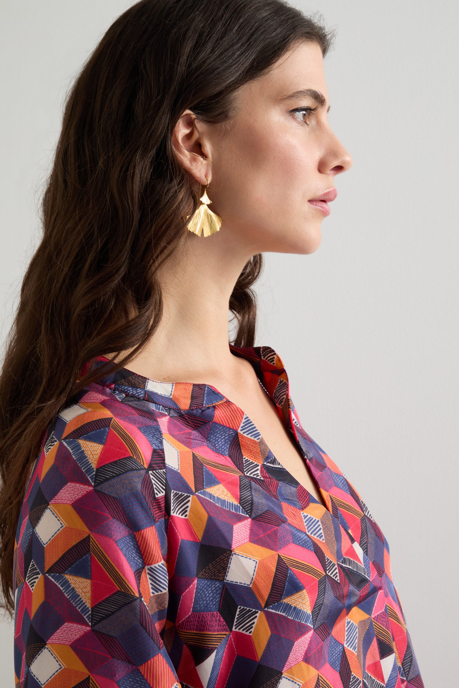 Woman with long hair in a Small Stitched Squares Silk Dress and gold fan-shaped earrings, looking to the side against a plain background.