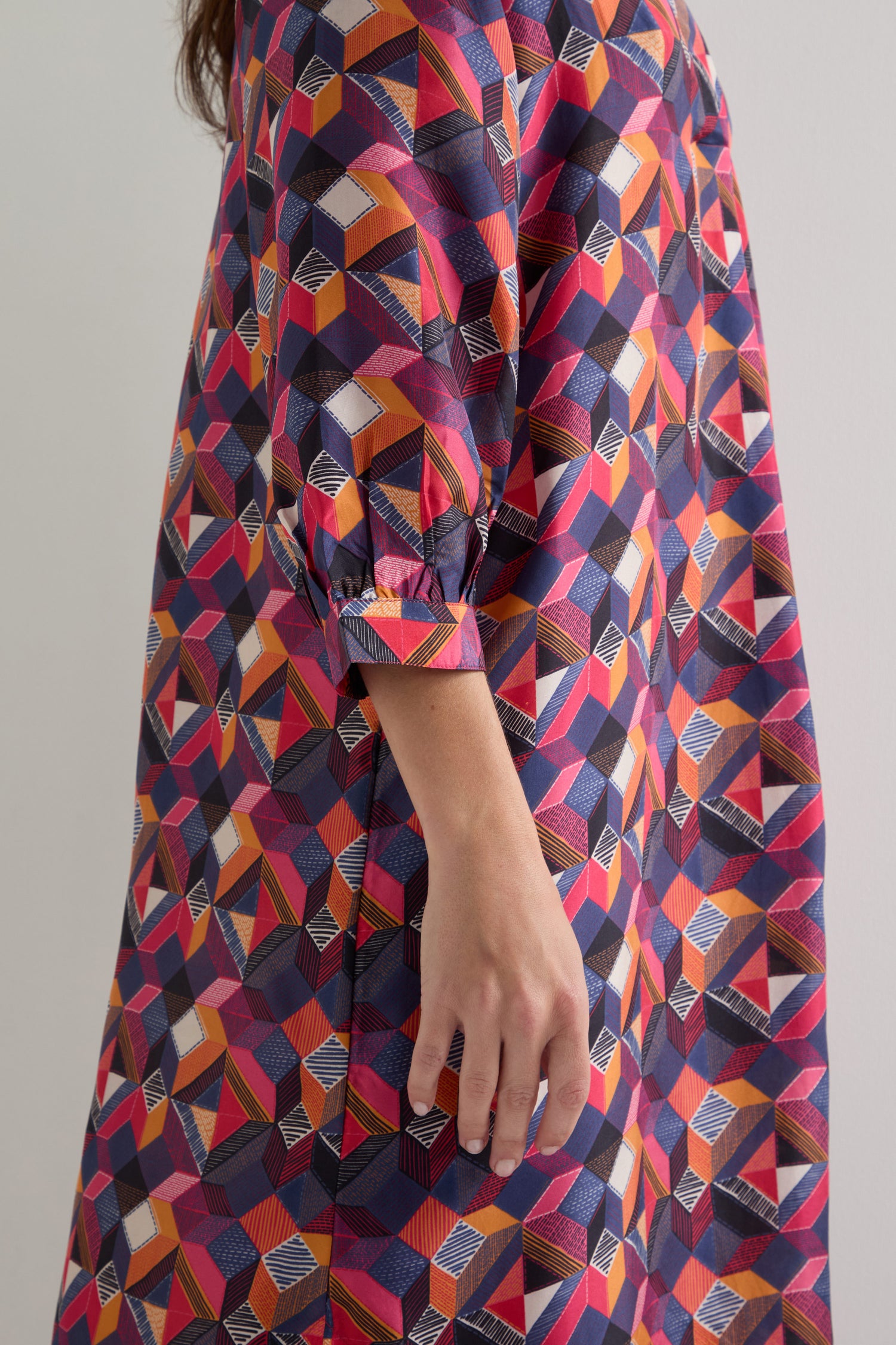 A person wearing the Small Stitched Squares Silk Dress, featuring a relaxed fit and vibrant geometric pattern, stands with their arm gently resting by their side.