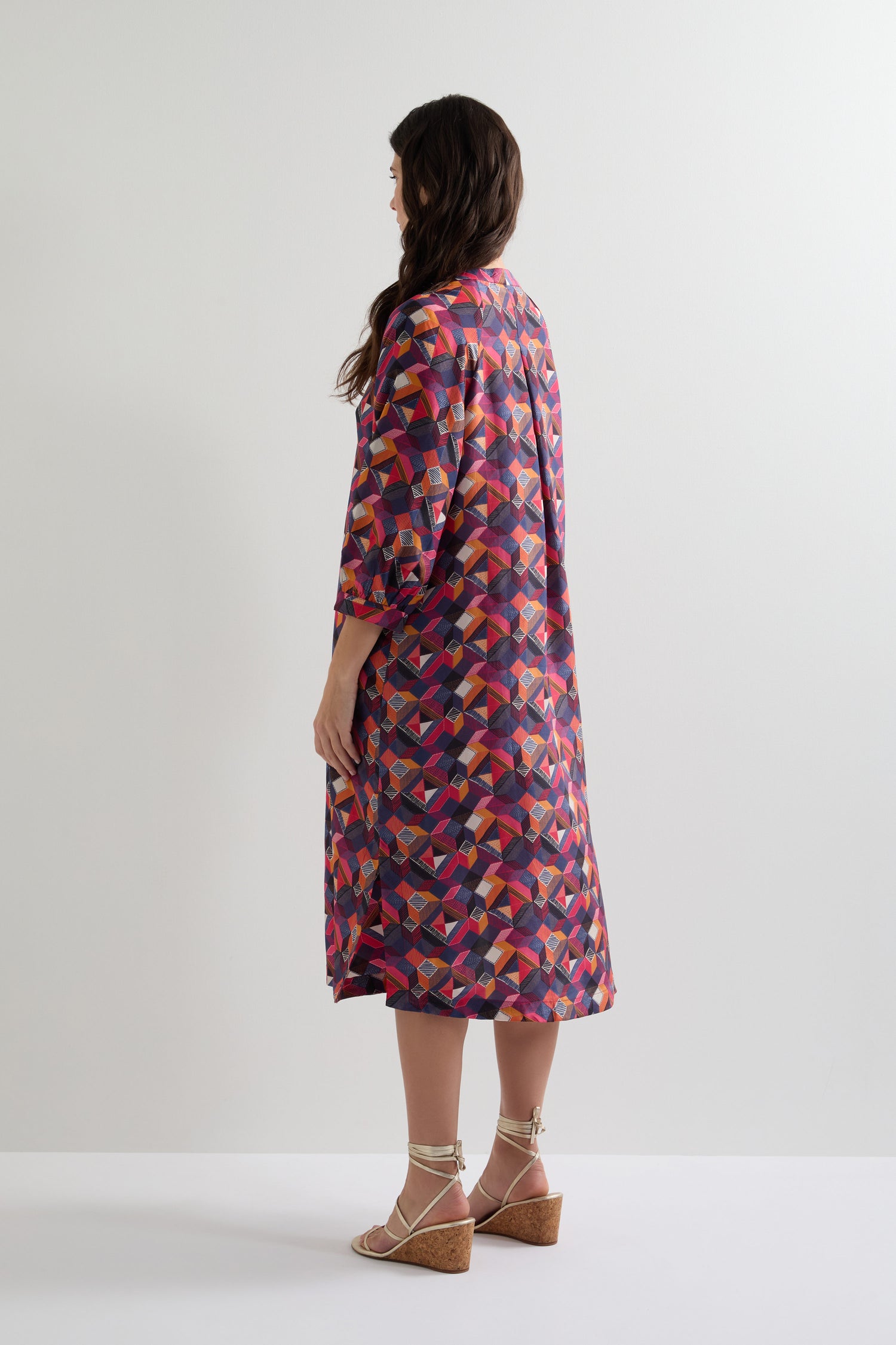 A person in a Small Stitched Squares Silk Dress, featuring a colorful geometric print and relaxed fit, wears wedge sandals while standing against a plain white background, slightly turned away from the camera.