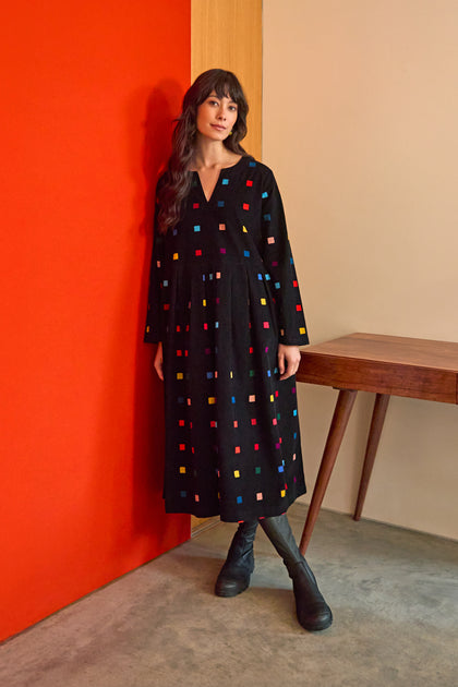 A woman in the Multi Colour Check Cord Print Dress stands by a wooden table against a red and beige wall on a concrete floor.