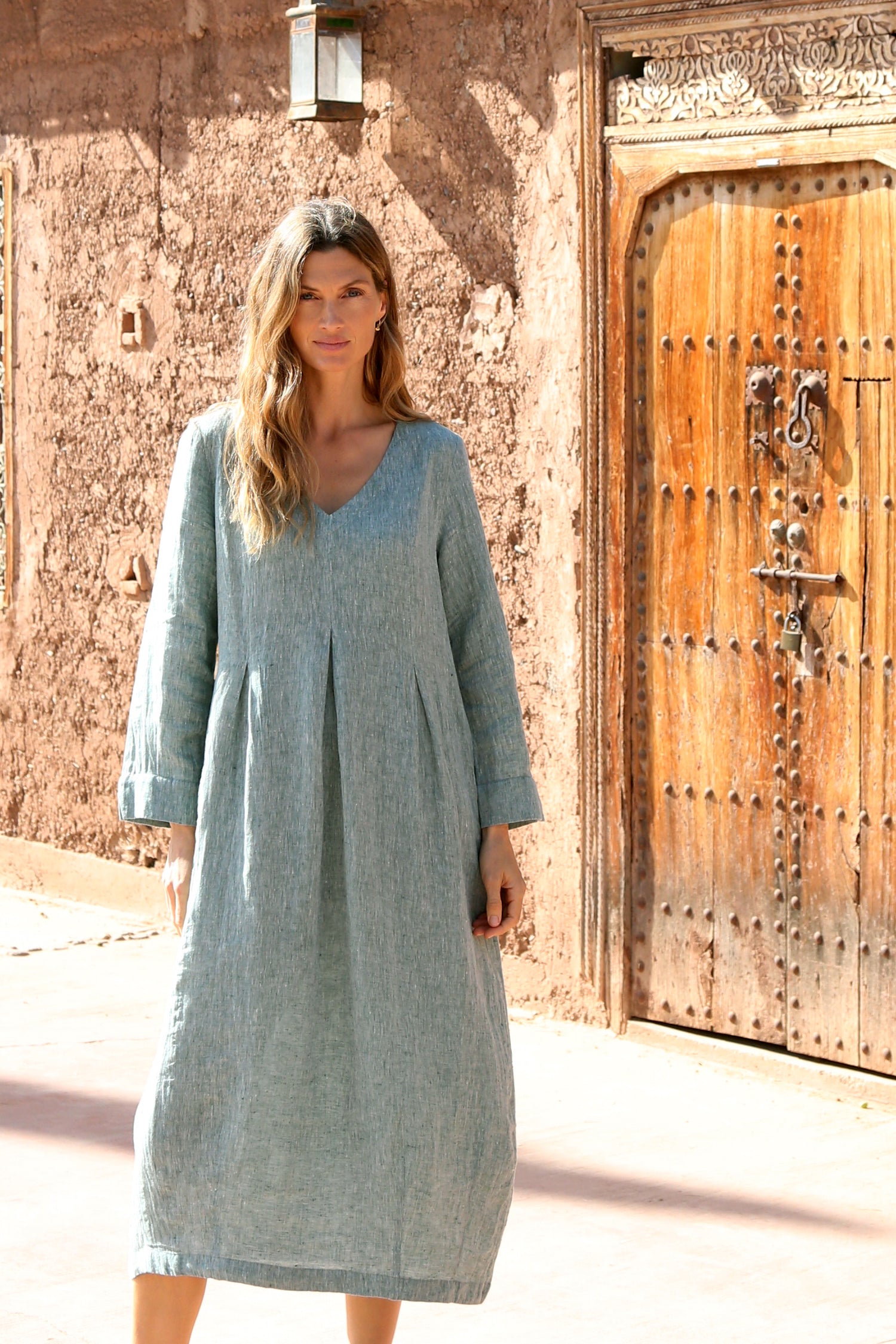 A woman in a loose-fitting, long-sleeved, light blue Crinkle Linen Cross Dye Bubble Dress stands before a rustic brown wall and ornate wooden door.