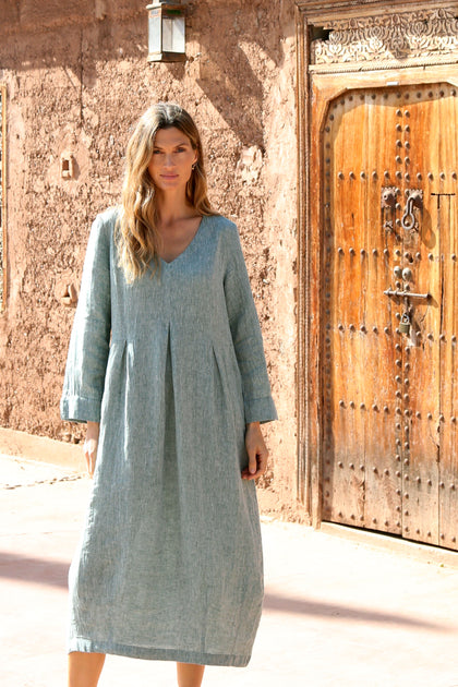 A woman in a loose-fitting, long-sleeved, light blue Crinkle Linen Cross Dye Bubble Dress stands before a rustic brown wall and ornate wooden door.