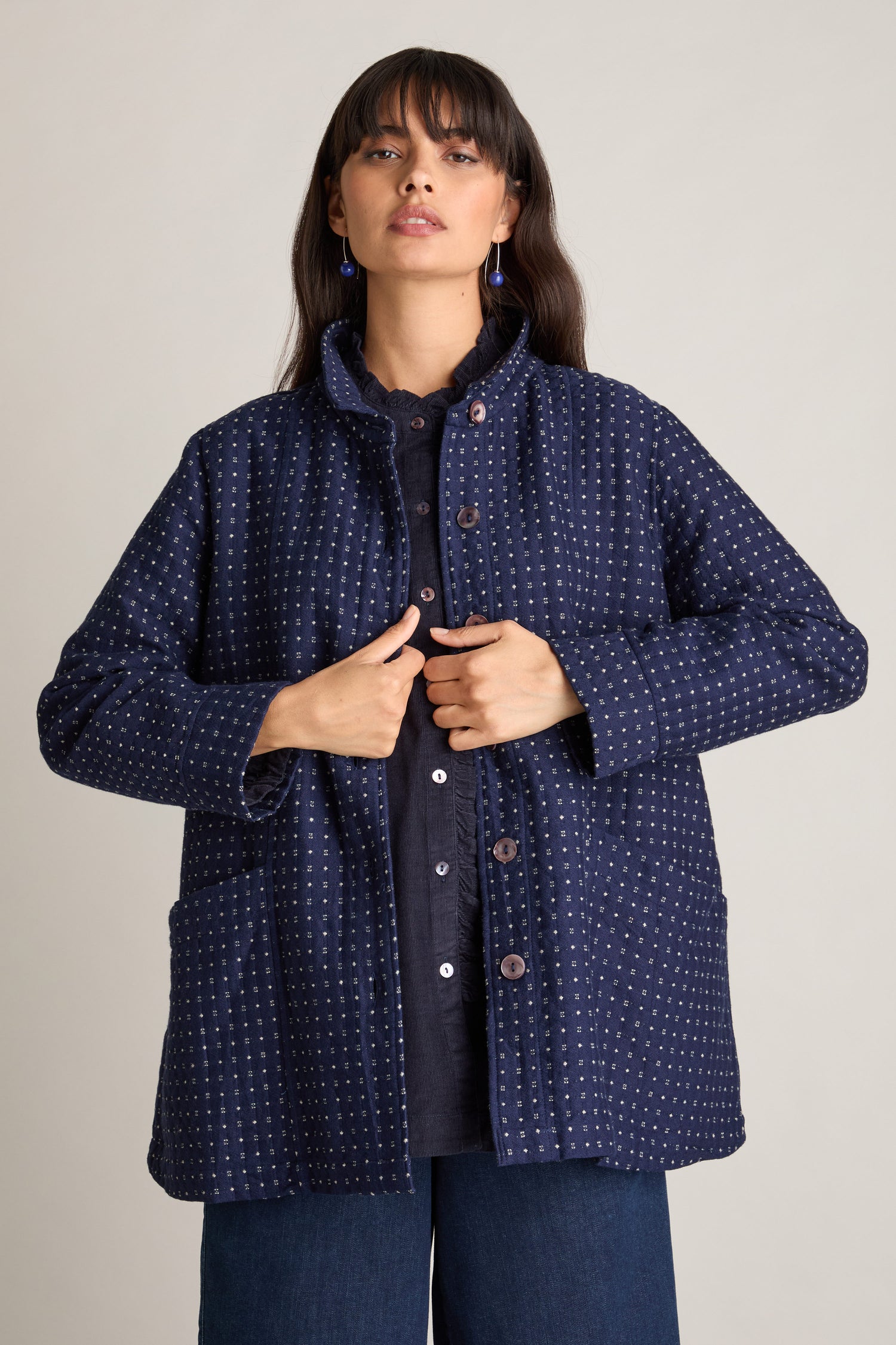 A woman stands against a neutral background wearing an Indigo Stitch Wool Cotton Jacket in a navy blue jacquard weave over a dark shirt and paired with blue jeans. She is buttoning the jacket and looking ahead, showcasing an Autumnal style.