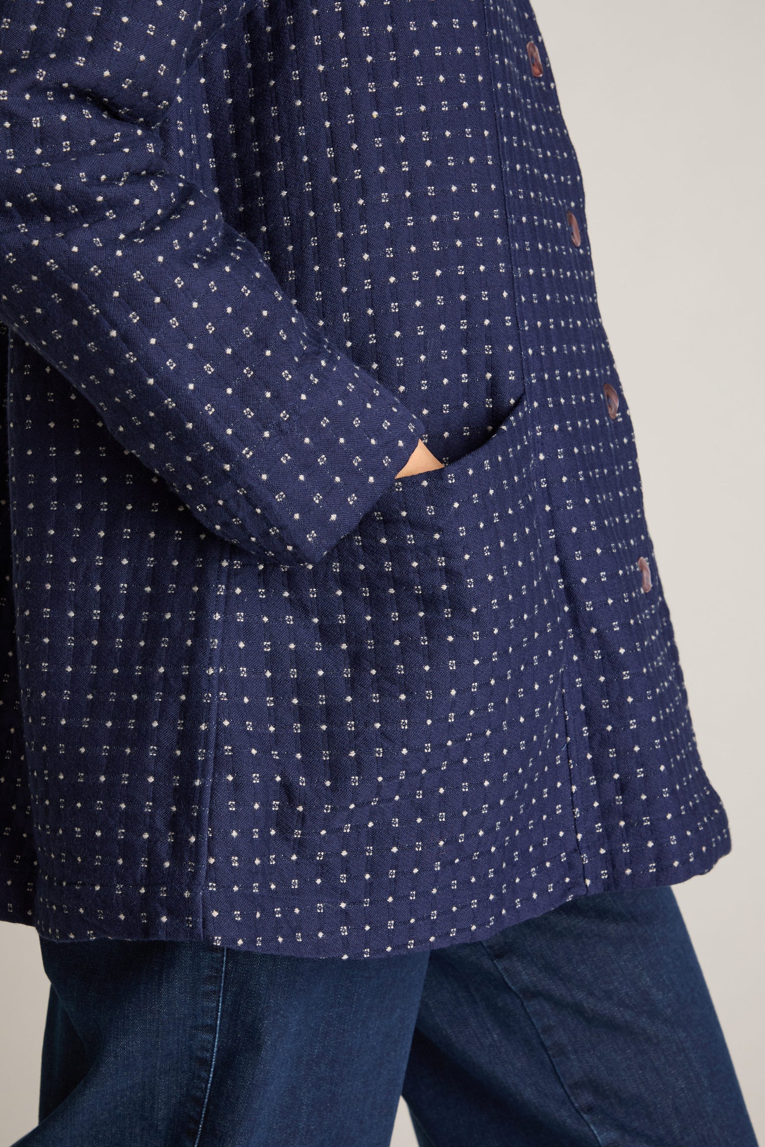 A person wearing the Indigo Stitch Wool Cotton Jacket with a blue jacquard weave and visible buttons has their hand partially tucked into the large front pocket, complementing their autumnal style. They are also wearing blue pants.