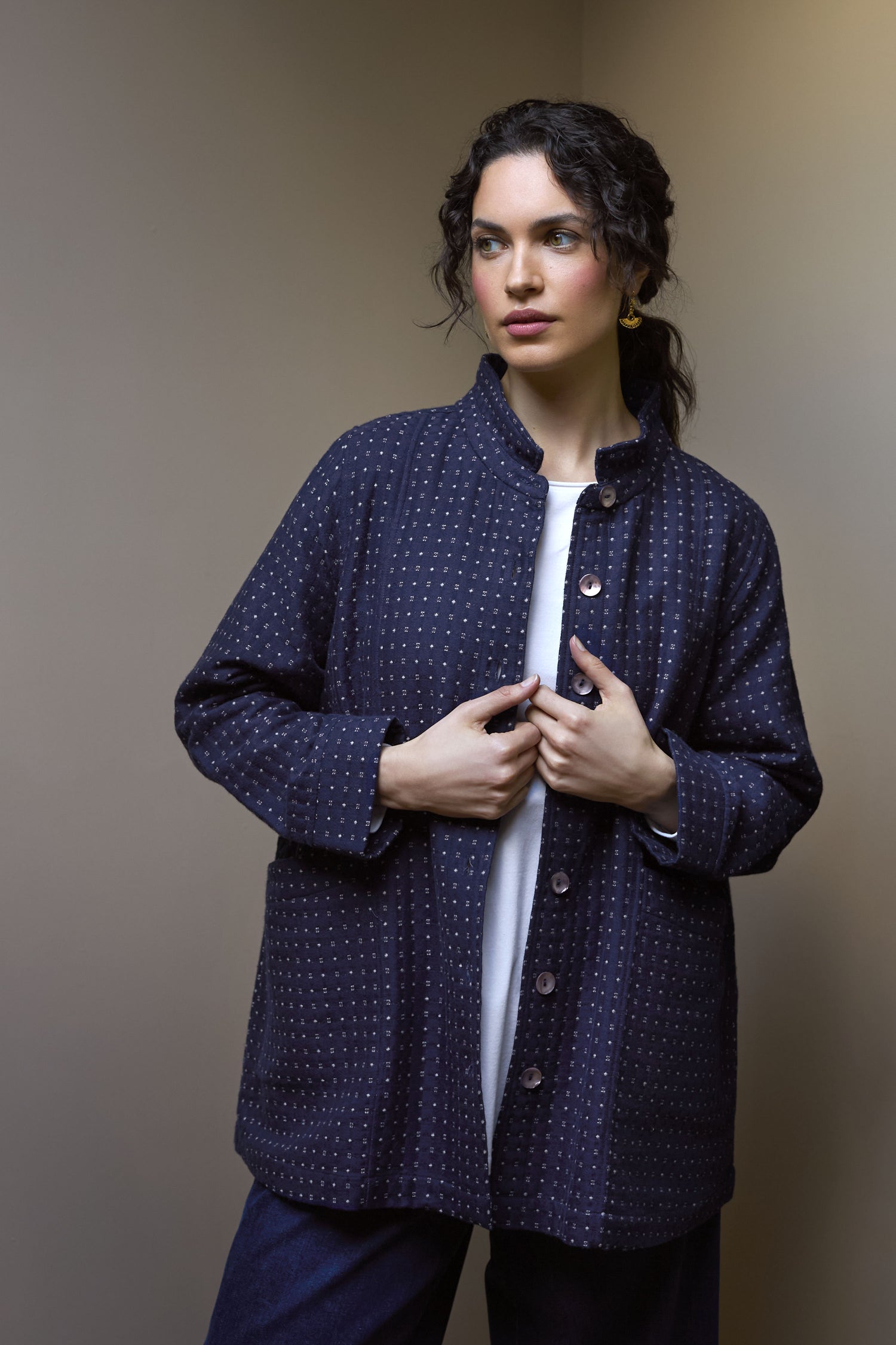 A woman stands against a plain background, wearing the Indigo Stitch Wool Cotton Jacket over a white shirt, with her hands adjusting the jacket's front.