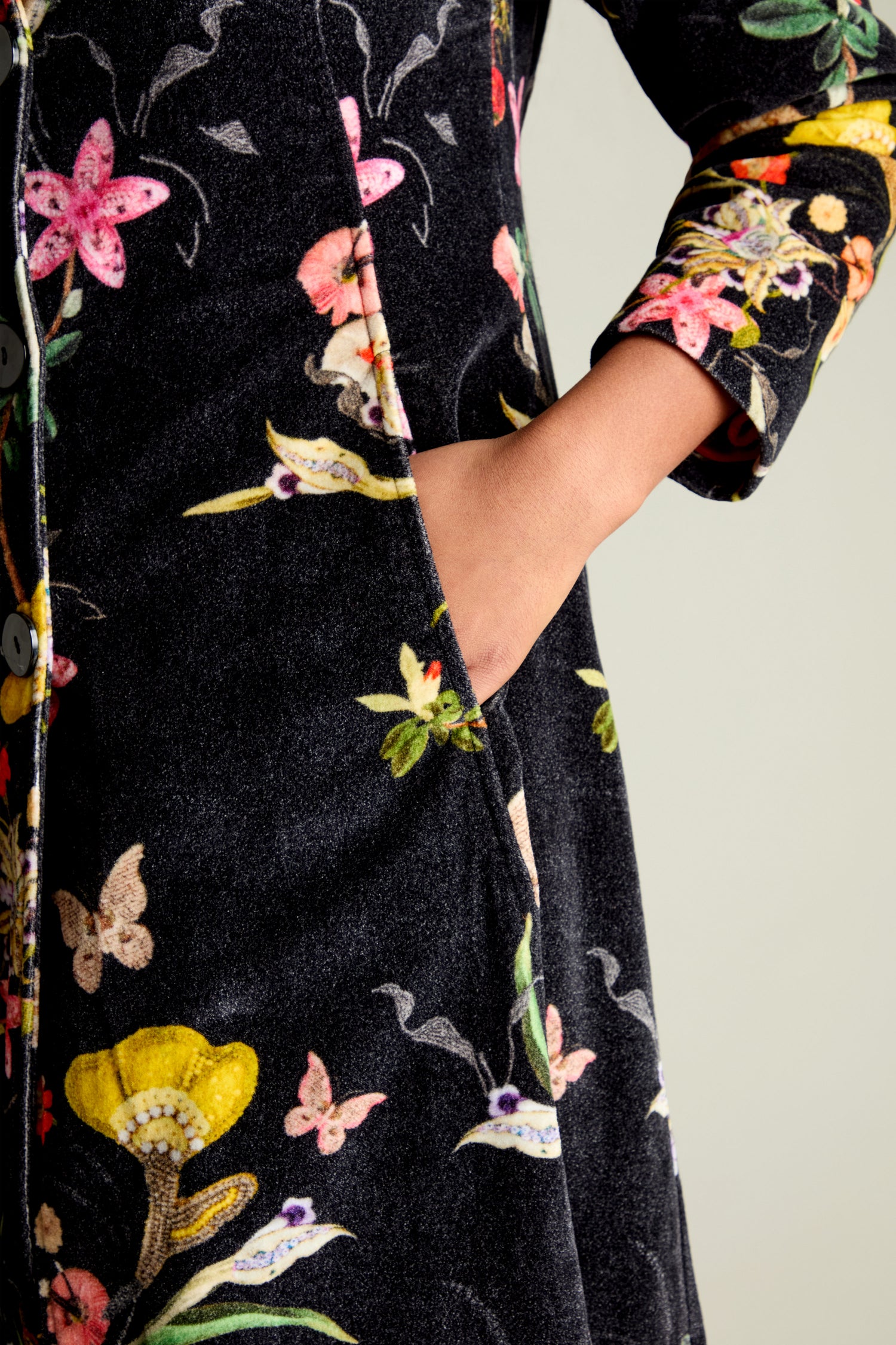 A person's hand rests in the pocket of this Velvet Vintage Floral Coat, featuring long sleeves adorned with delicate flowers and butterflies on a light background, complemented by an elegant shawl collar.