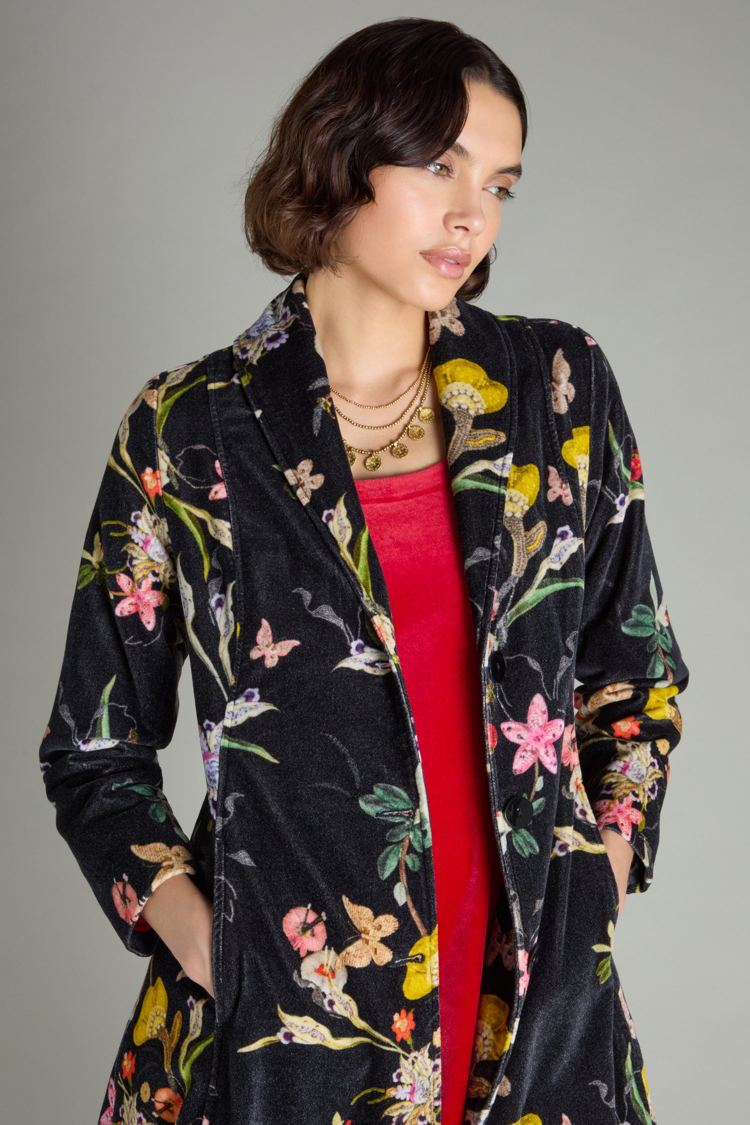 A person is posing against a plain background, wearing the Velvet Vintage Floral Coat with a shawl collar over a red top.