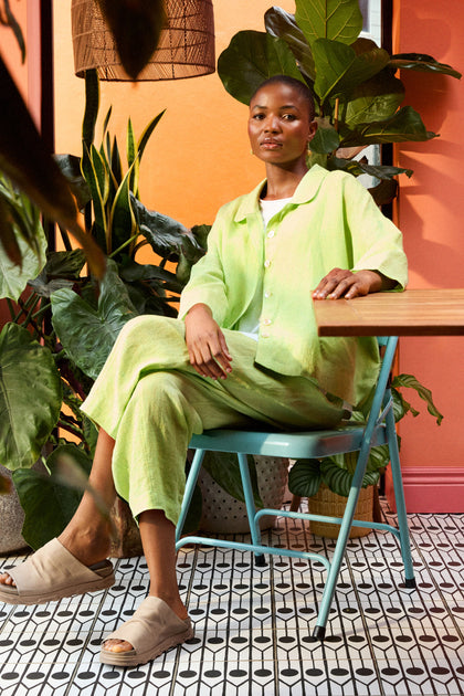 A person in a Linen Ticking Stripe Boxy Jacket sits on a teal chair, surrounded by plants against an orange wall and patterned floor.