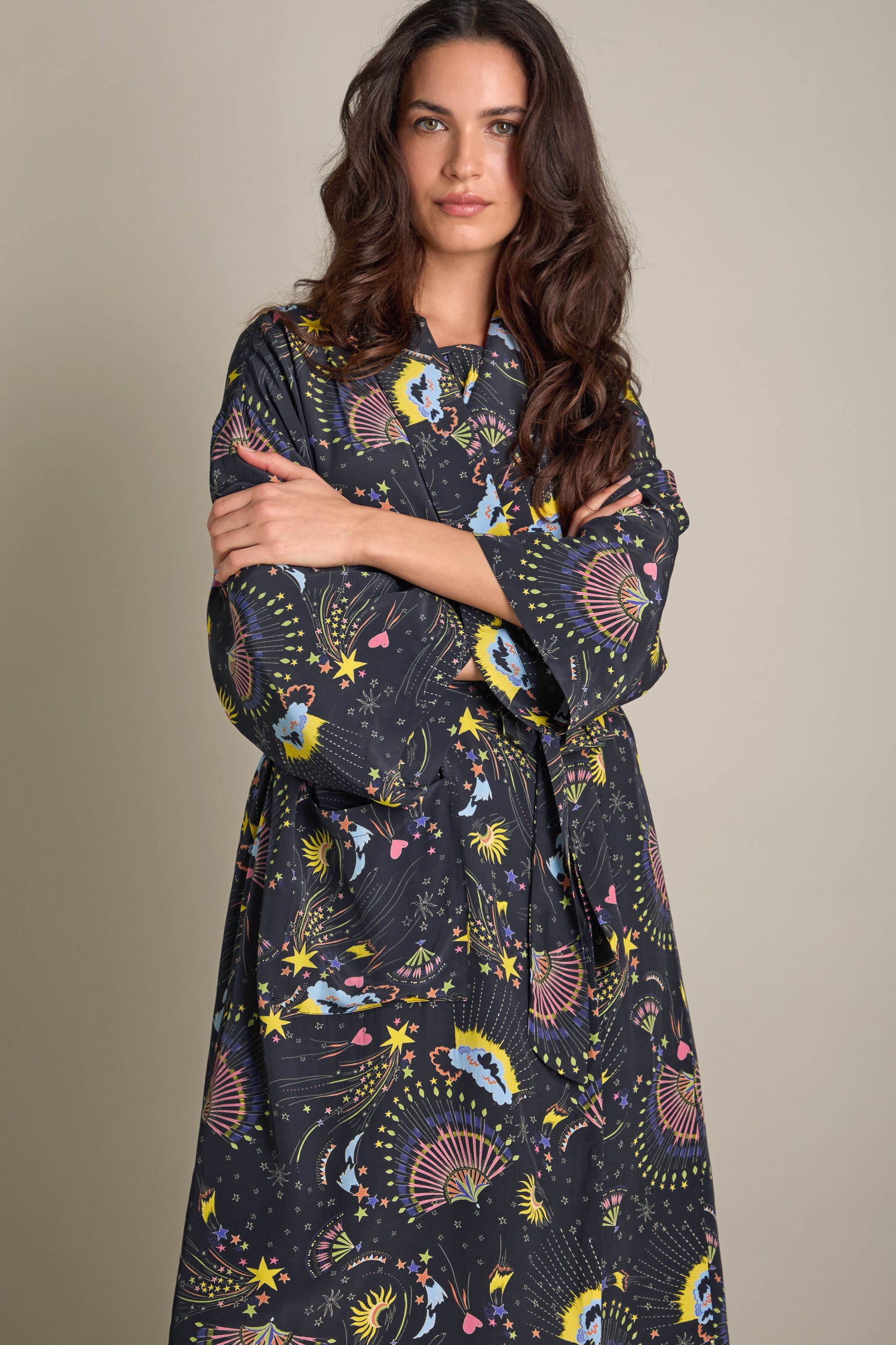 Wearing the Celestial Dreams Robe, a person stands with arms crossed against a plain background, exuding an air of elegant design with its silky texture and intricate patterns.