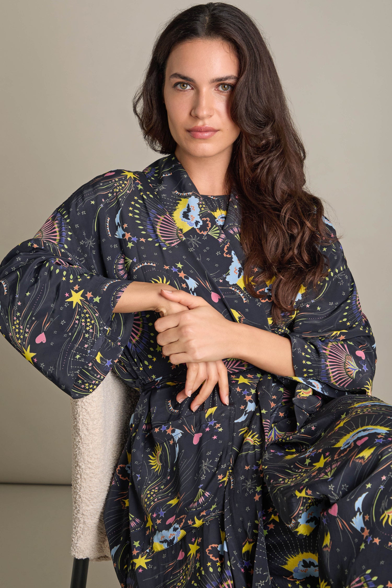 A woman with long dark hair sits on a light-colored chair, wrapped in the Celestial Dreams Robe adorned with colorful designs. The shawl collar adds an element of luxurious comfort as she rests her hands gracefully crossed in front of her against the plain background.