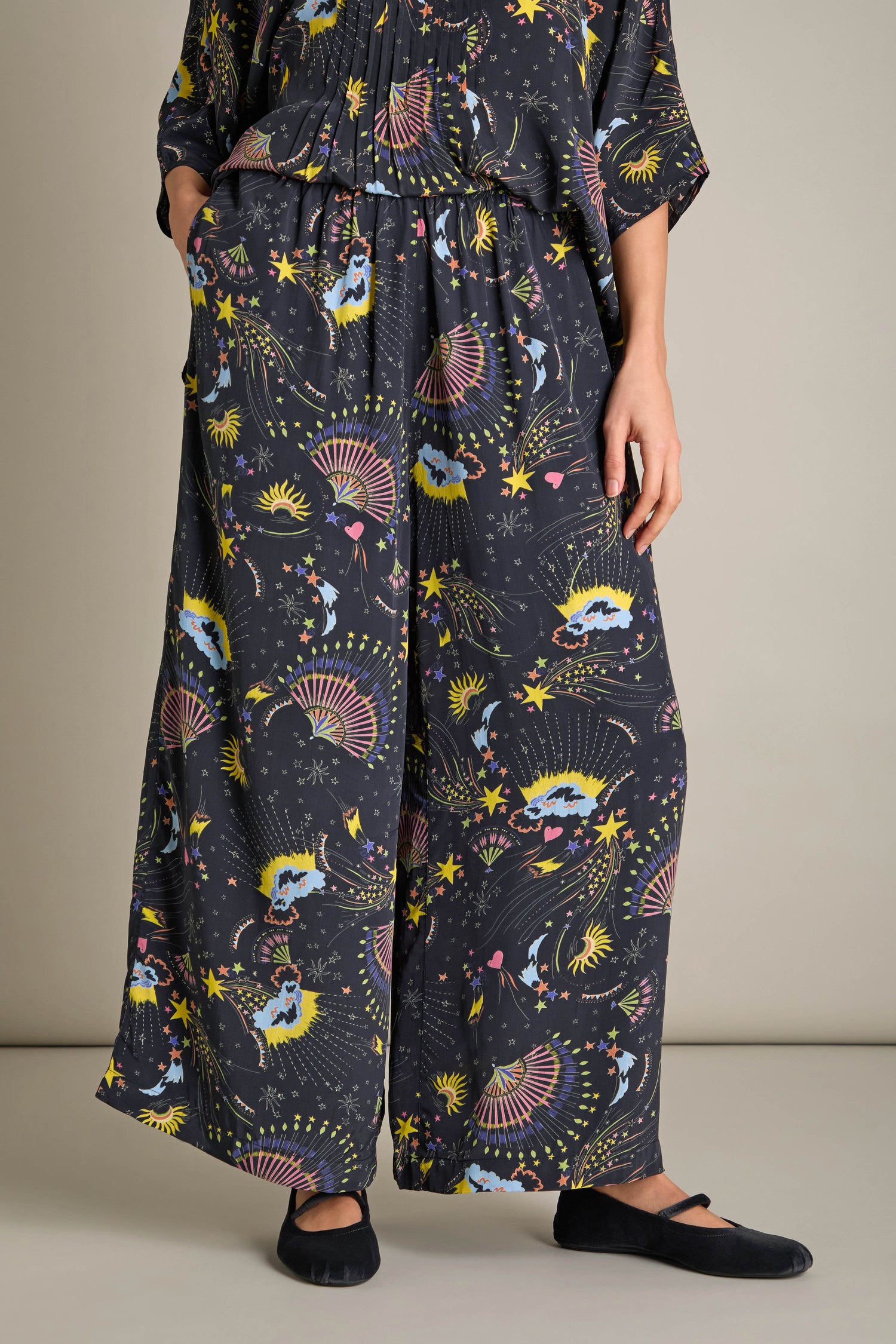 A person stands gracefully against a neutral background, wearing the Celestial Dreams Pyjama Set, an elegant dark patterned outfit featuring wide-leg pants and black shoes.