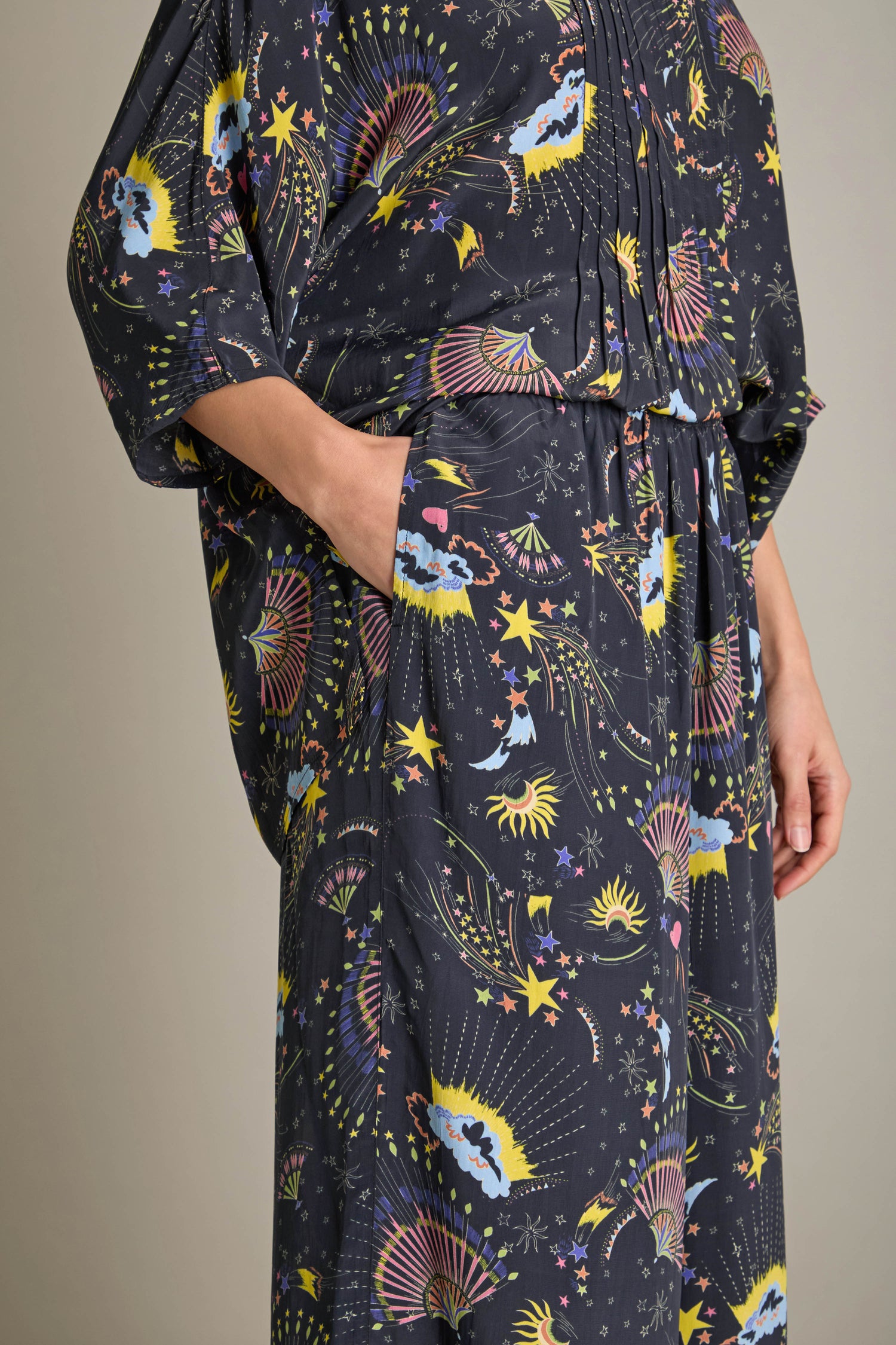 Person dressed in the Celestial Dreams Pyjama Set, featuring a dark fabric adorned with vibrant abstract patterns of stars and sunbursts. The relaxed fit of this dream-inspired piece provides comfort, allowing them to easily slip one hand into a pocket.