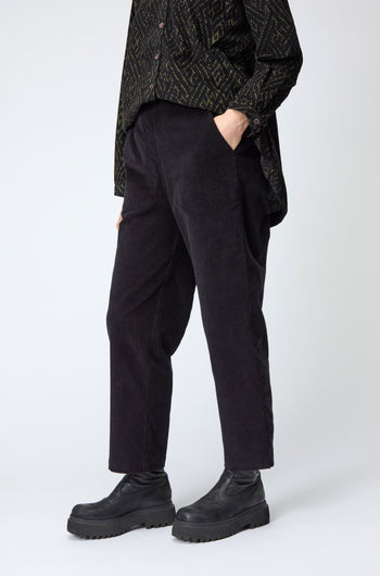 Person wearing Cord Straight Leg Black Trousers, a black patterned shirt, and black platform shoes, stands against a plain light background.