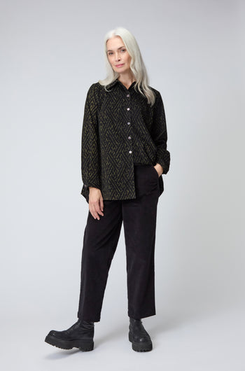 A woman with long gray hair stands facing forward, wearing a patterned button-up shirt, Cord Straight Leg Black Trousers, and black boots against a plain light background.