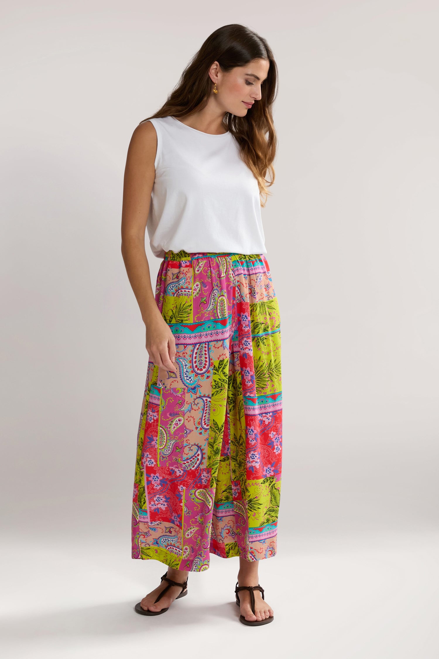 A woman in a white sleeveless top and Tropical Patchwork Wide Trouser stands against a plain background, looking down and slightly to the side. She is wearing black sandals.