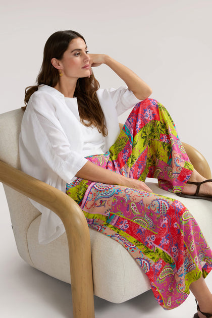 A person with long hair sits on a beige chair, wearing a white blouse and Tropical Patchwork Wide Trouser, looking to the side with one hand resting on their head.