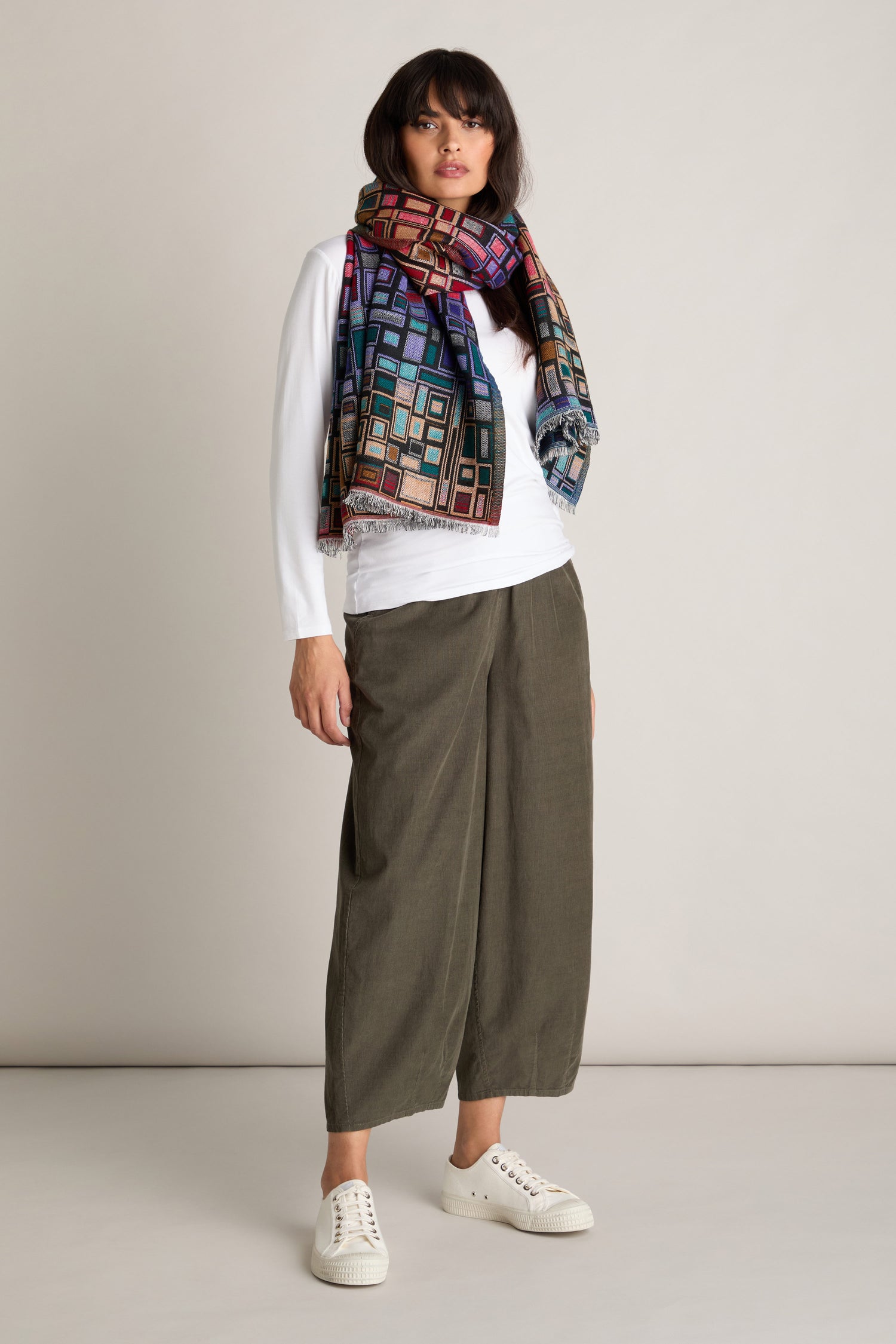 A person stands against a plain wall wearing a white long-sleeve shirt, olive green Pinwale Cord Bubble Trousers with an elasticated waist, white sneakers, and a colorful geometric-patterned scarf.