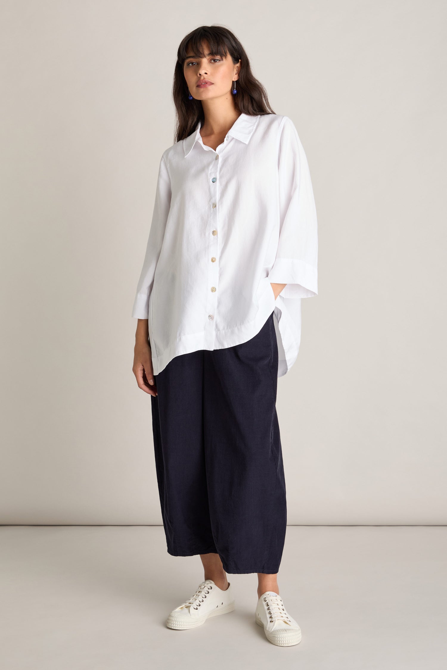 A person stands wearing a white button-down shirt, dark loose-fitting Pinwale Cord Bubble Trousers with an elasticated waist, and white sneakers against a plain background.