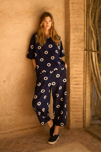 A woman stands against a beige textured wall, wearing Ikat Circles Cotton Bubble Trousers—loose-fitting navy pants with a white circular pattern—paired with a matching top and black slip-on shoes.
