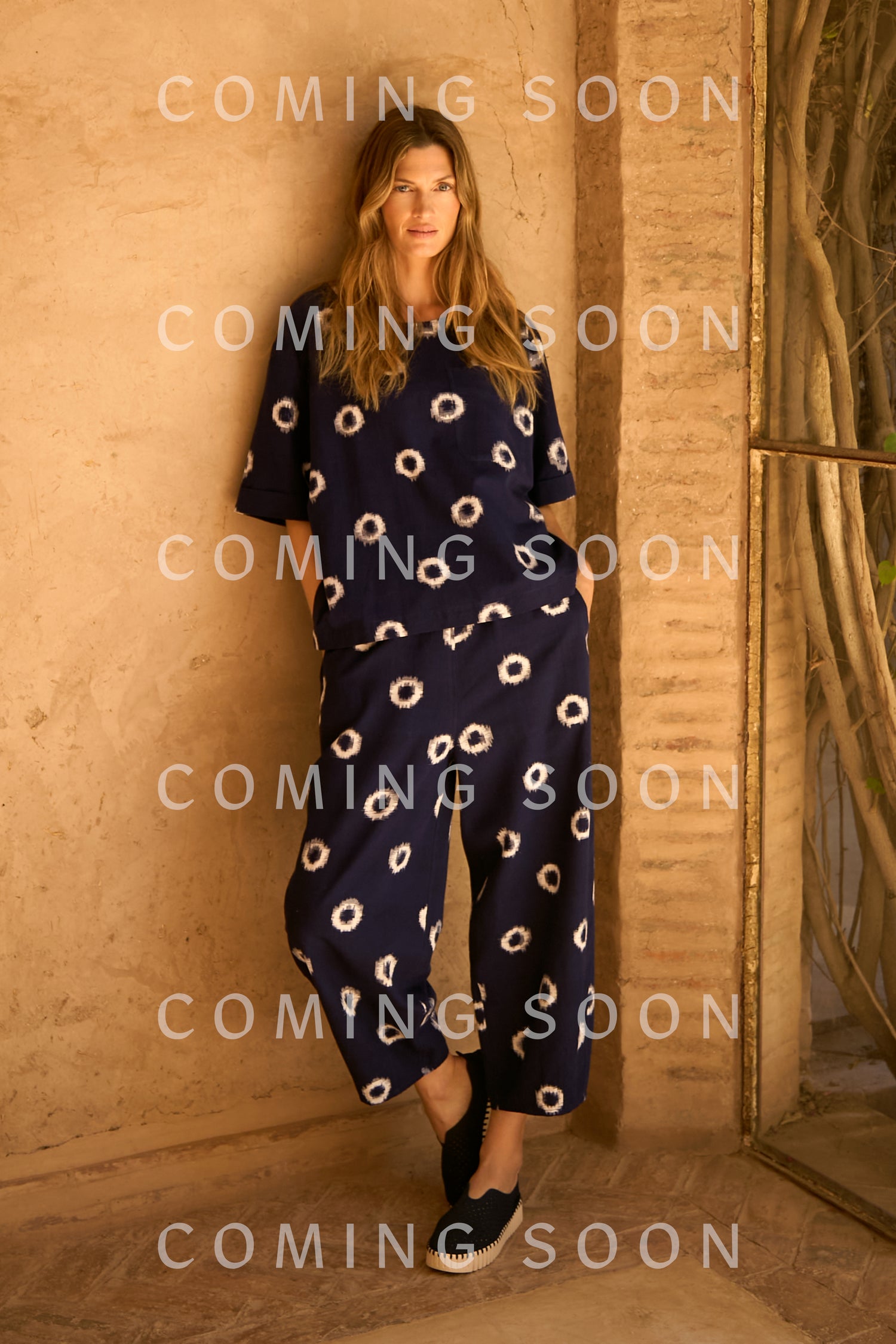 A woman stands against a tan stucco wall in Ikat Circles Cotton Bubble Trousers, a loose-fitting navy style with white circle patterns and black slip-on shoes; "COMING SOON" text is overlaid multiple times.
