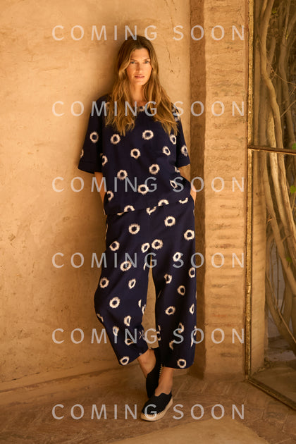 A woman stands against a tan stucco wall in Ikat Circles Cotton Bubble Trousers, a loose-fitting navy style with white circle patterns and black slip-on shoes; "COMING SOON" text is overlaid multiple times.