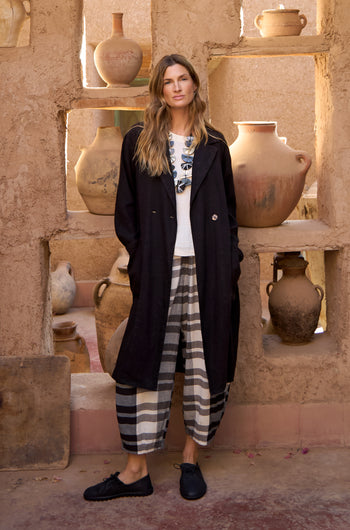 A woman stands by a rustic wall with pottery, modeling the GRP5167 look: a black coat, white top, Monochrome Crinkle Check Bubble Trouser, and black shoes.