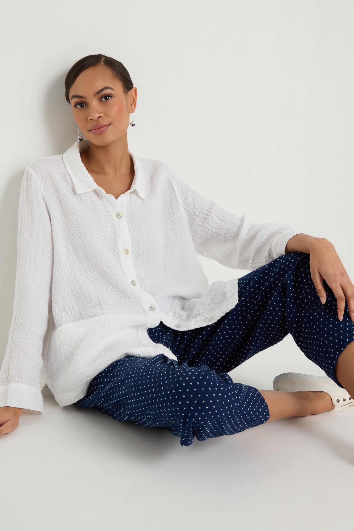 A person sits against a white wall wearing a textured white button-up shirt, Indigo Spot Cotton Bubble Trouser with an elasticated waist and navy blue polka dot pattern, paired with white sneakers.
