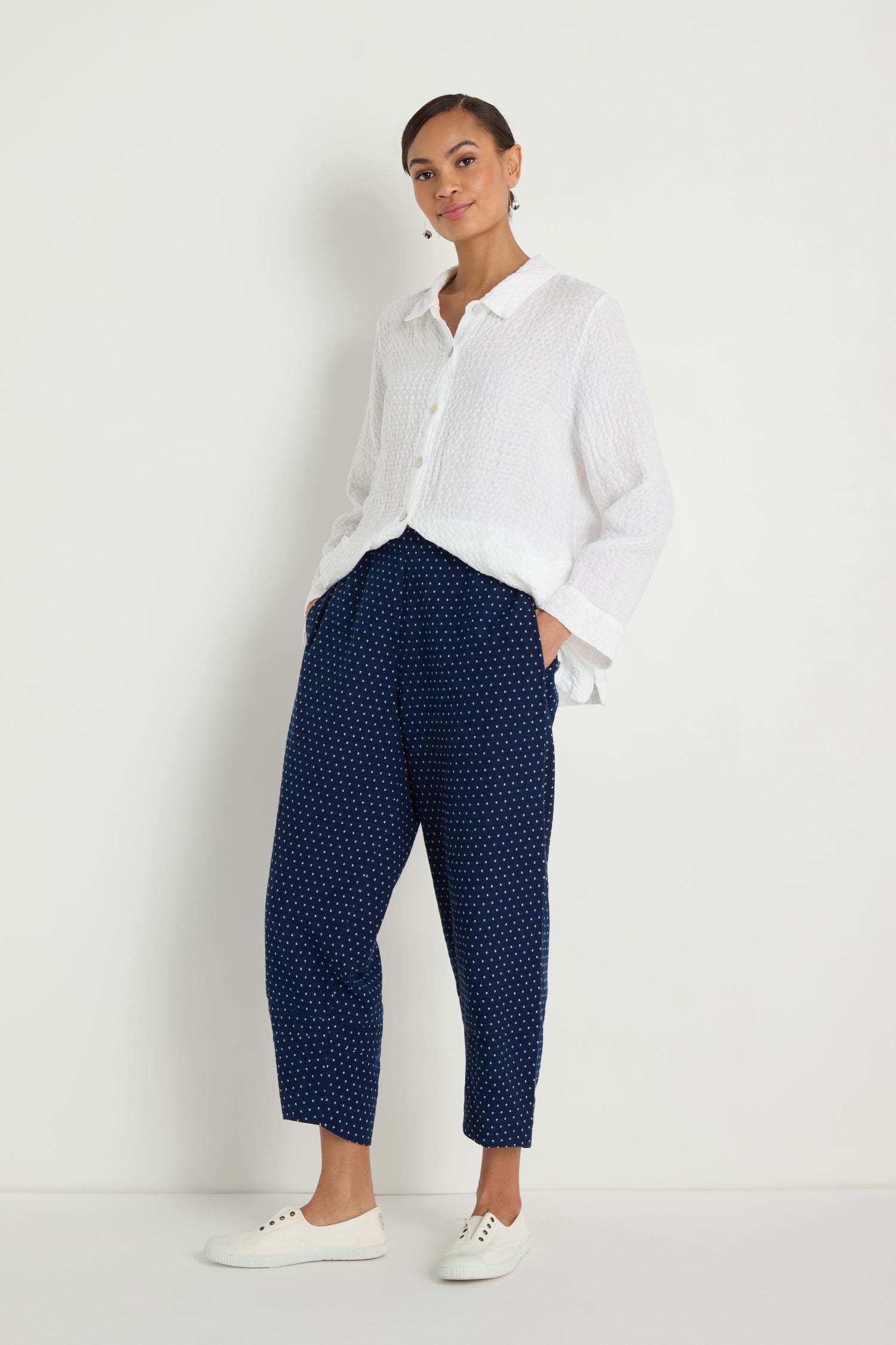 A woman stands against a white wall wearing a white button-up shirt, Indigo Spot Cotton Bubble Trouser with an elasticated waist, and white sneakers, hands in her pockets.