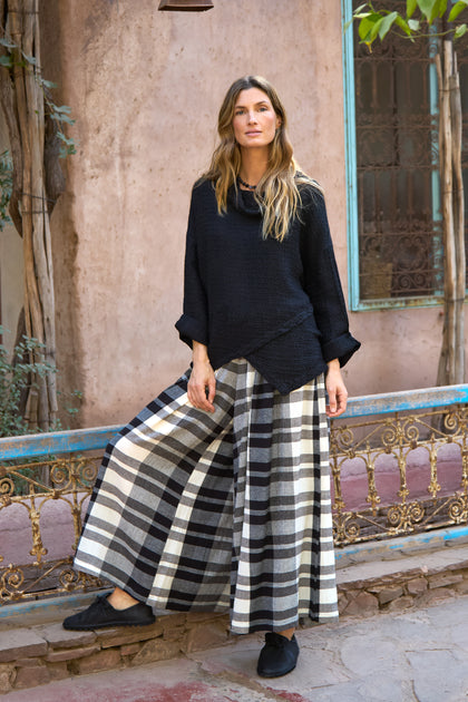 A woman stands outdoors beside a rustic metal railing, wearing Monochrome Crinkle Check Wide Trousers and a black long-sleeve top, with plants and a weathered wall in the background.