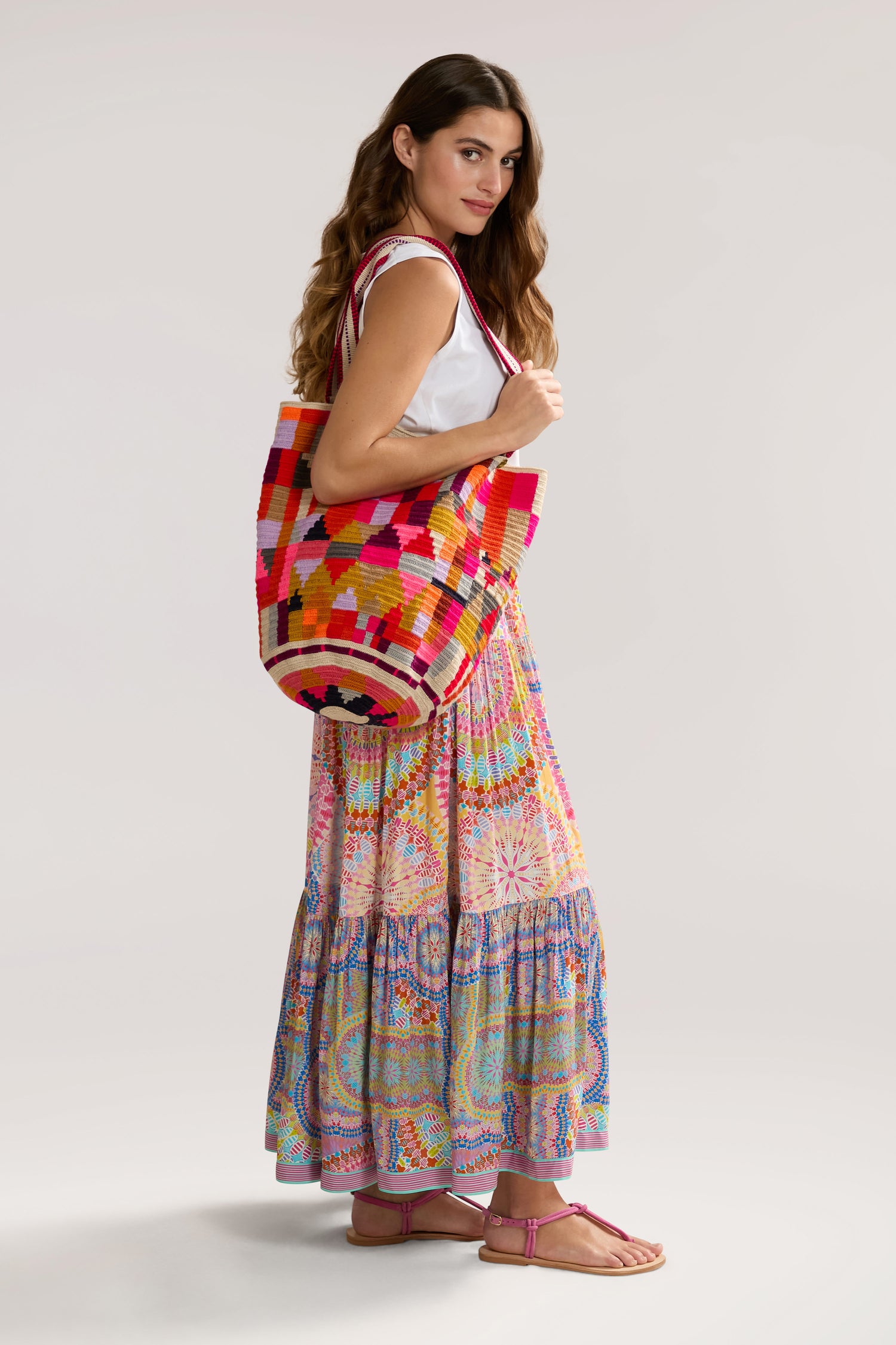 A woman with long hair is wearing a sleeveless white top and a multicolored Circle Mandala Print Skirt with an elasticated waist, holding a large colorful woven bag. She is standing sideways, exuding bohemian luxury.