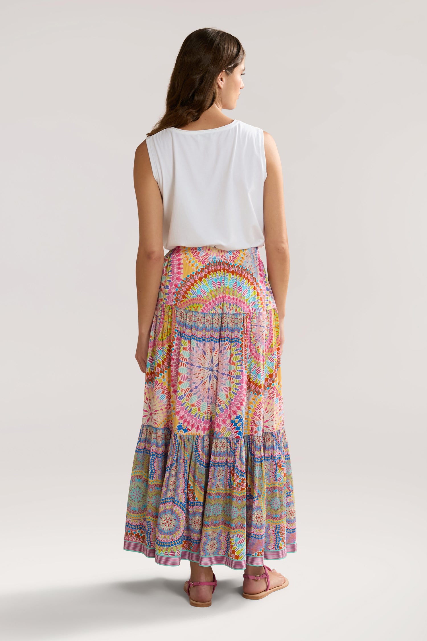 A woman with long hair, wearing a white sleeveless top and a colorful Circle Mandala Print Skirt with an elasticated waist, stands facing away from the camera against a plain background.