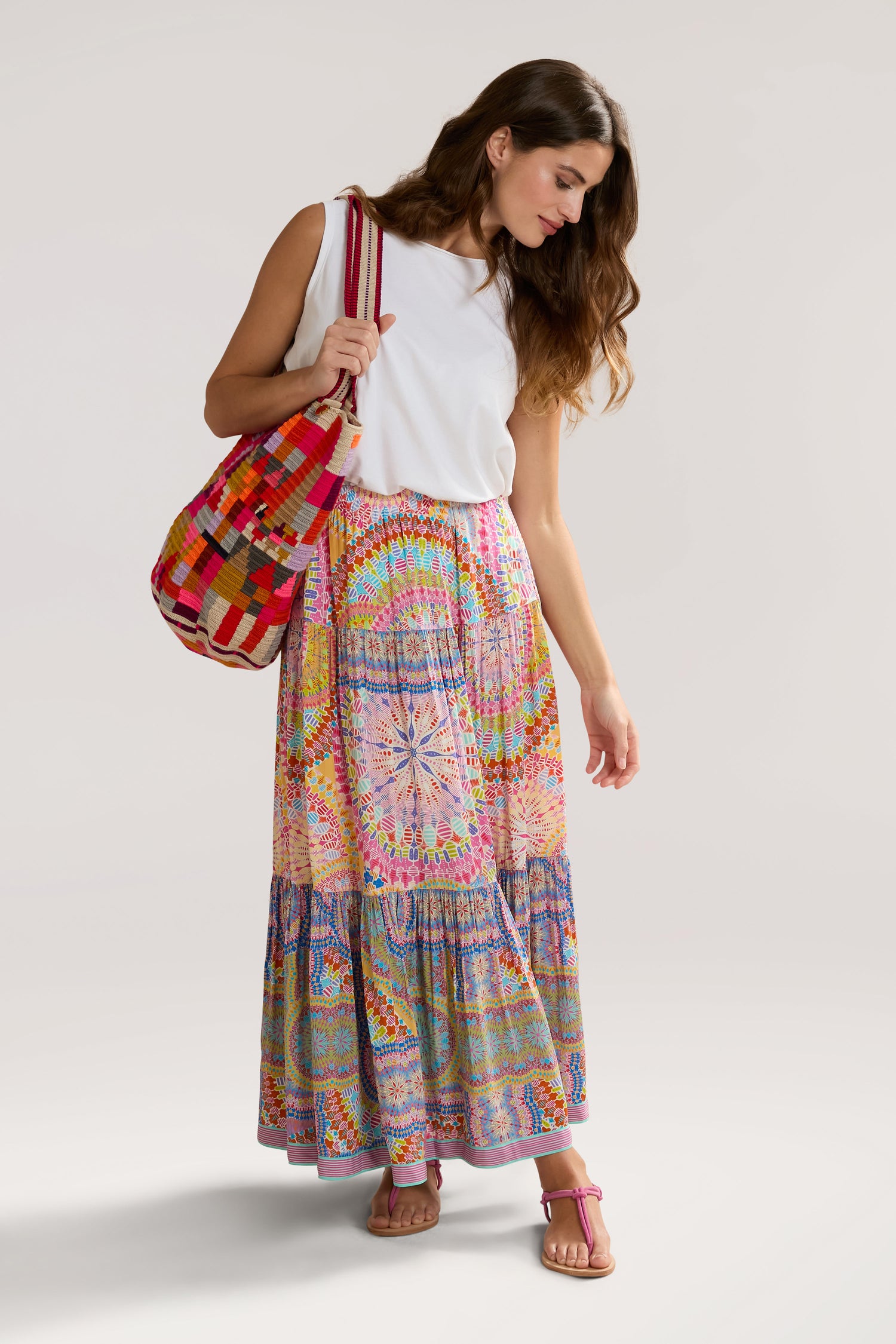 A woman in a white sleeveless top and a colorful, Circle Mandala Print Skirt with an elasticated waist holds a multicolored woven bag over her shoulder. She is looking down and has long, wavy hair. She wears pink sandals that complement her bohemian luxury style.