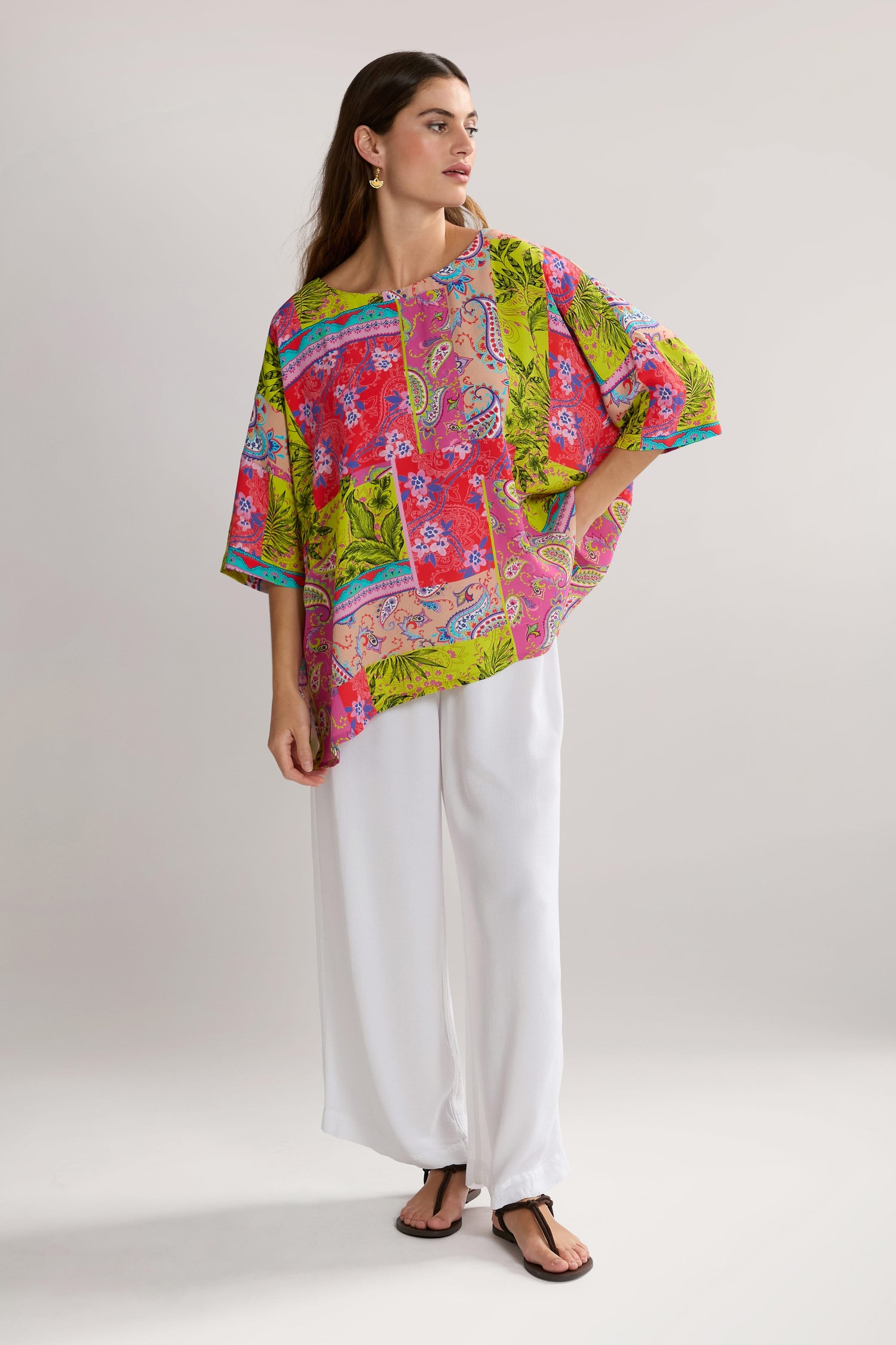 A person stands wearing a bright, multicolored Tropical Patchwork Relaxed Top in pure viscose and white pants, posing with one hand on their hip and looking slightly to the side, exuding a bohemian style.