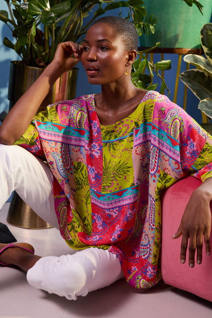 A person with short hair sits on the floor, wearing a Tropical Patchwork Relaxed Top and white pants. They are surrounded by plants and resting their elbow on a pink cushion.