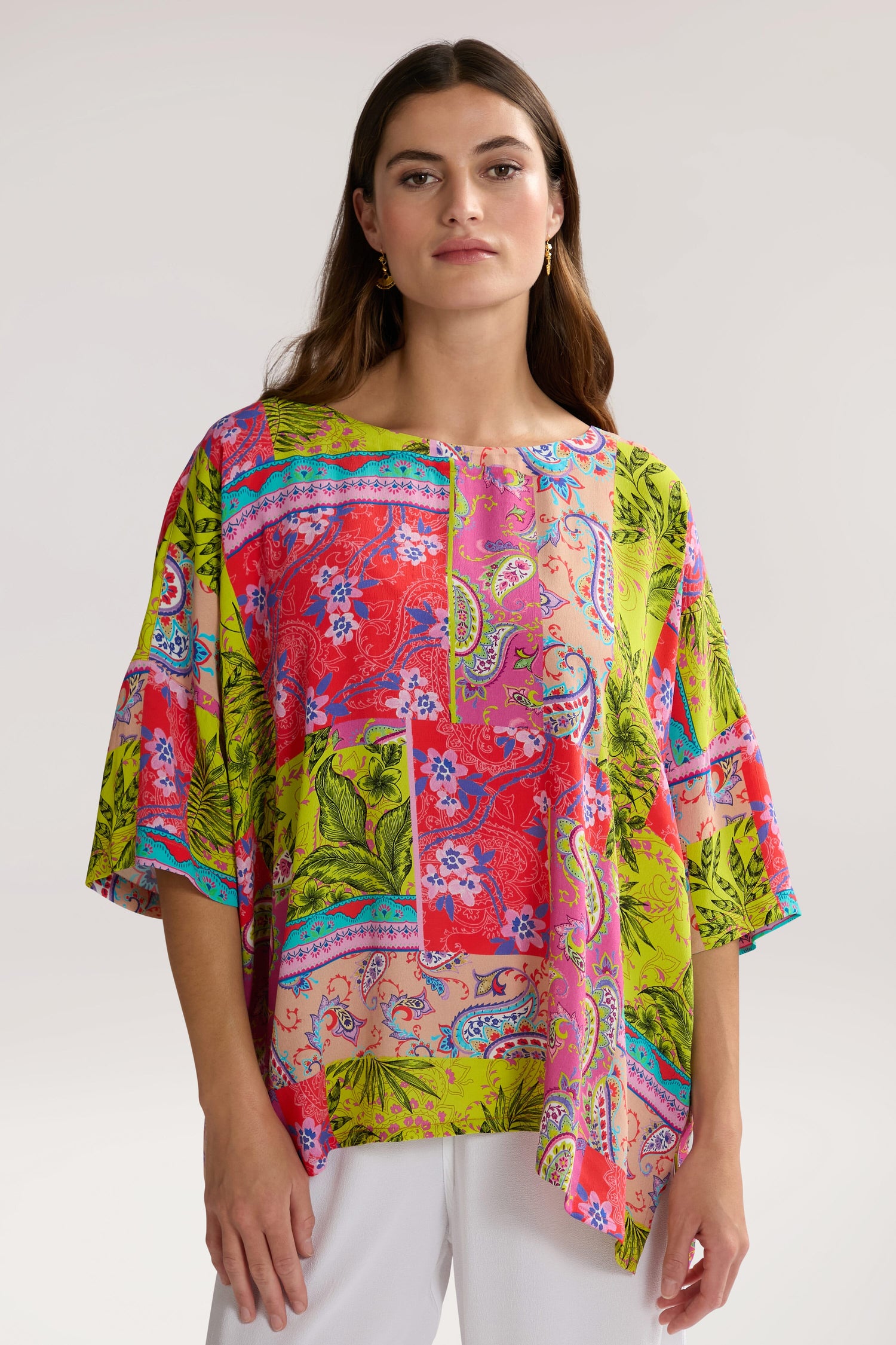 A woman is wearing a brightly colored Tropical Patchwork Relaxed Top with floral and abstract designs and white pants, posing against a plain background.