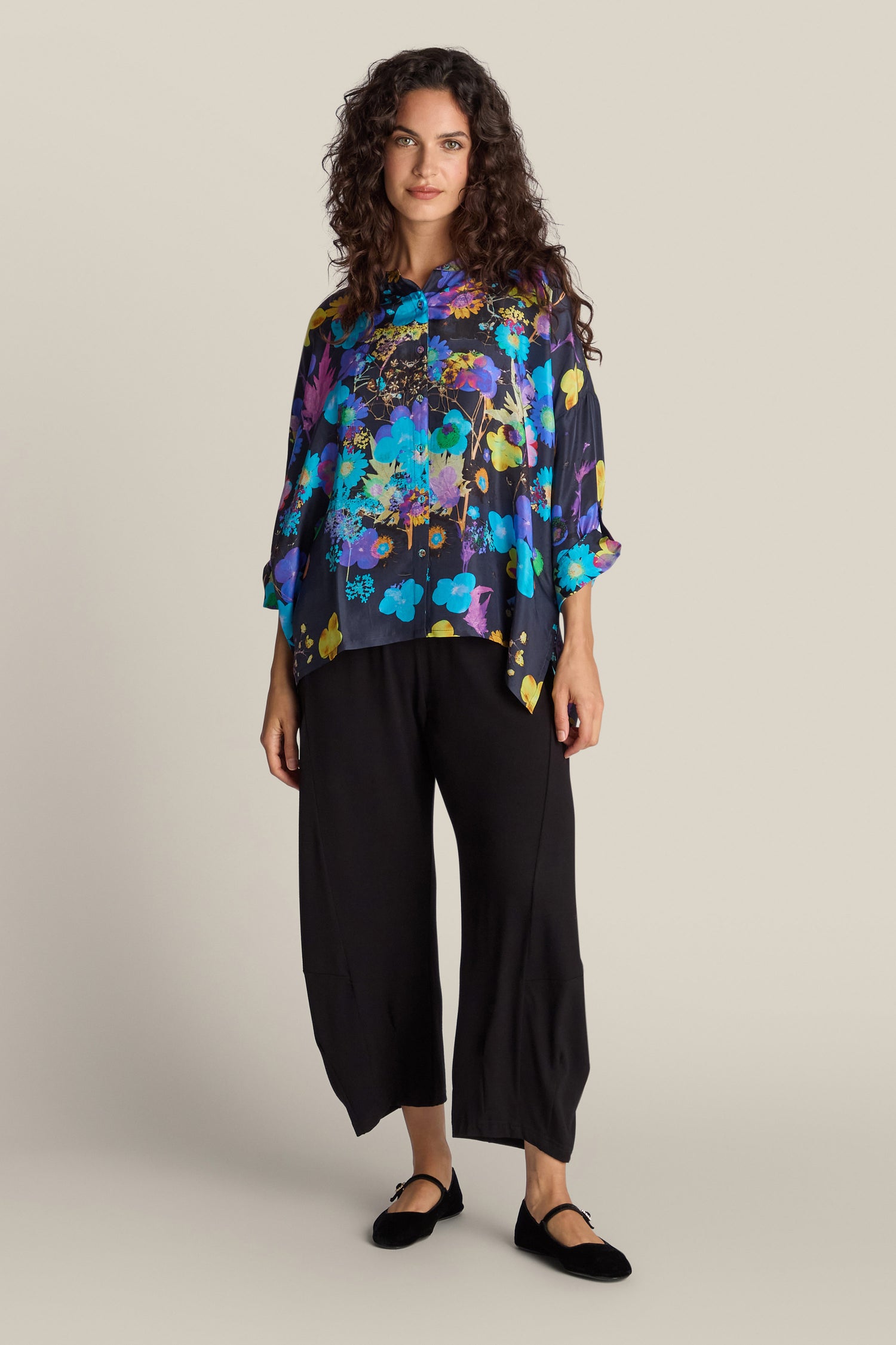 A person with curly hair in a vibrant Blue Bouquet Silk Shirt and loose black pants stands against a plain beige background, radiating floral flair. They are wearing black flats.